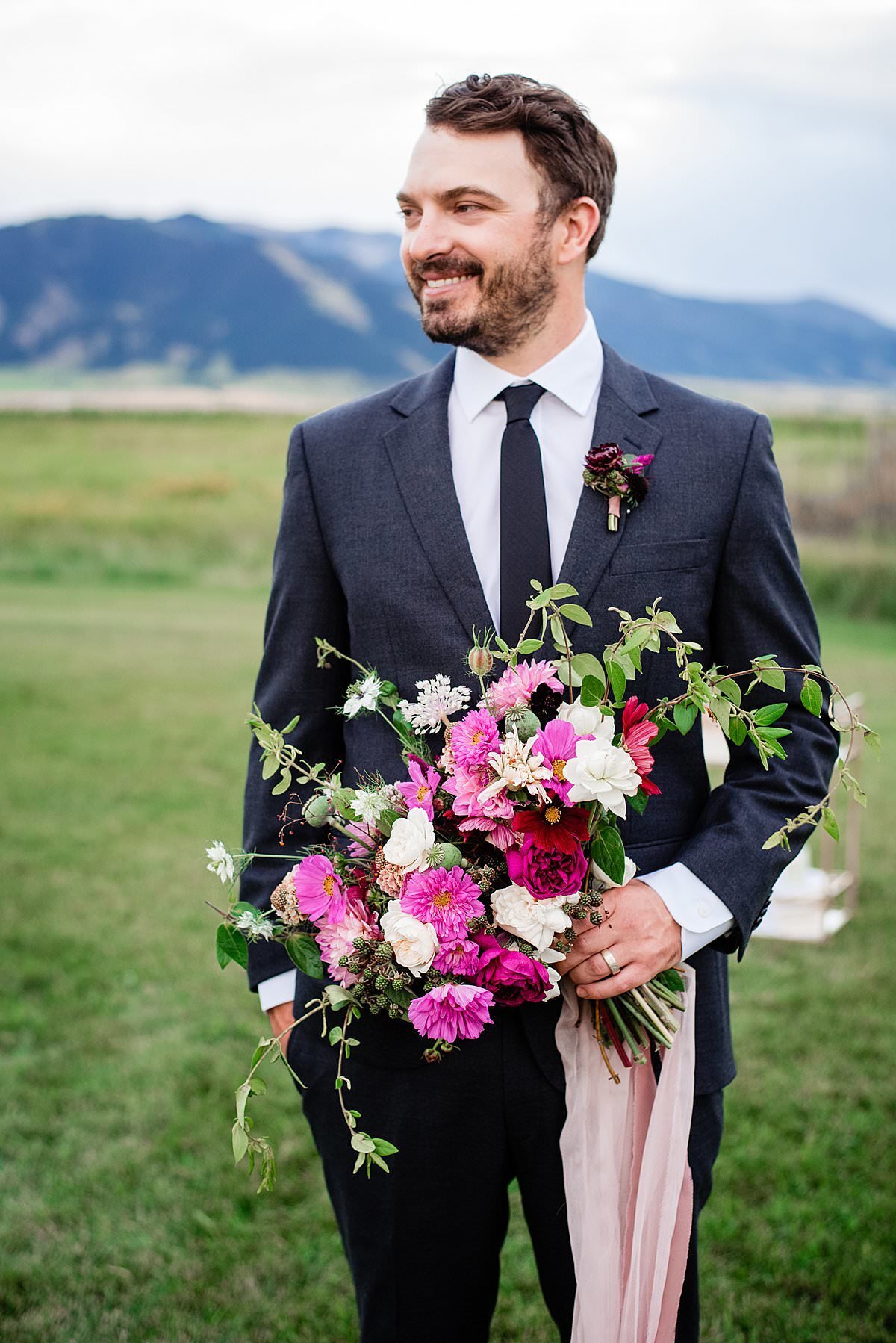 Groom holding large whimsical pink, white and burgundy bouquet wearing a sleek modern suit
