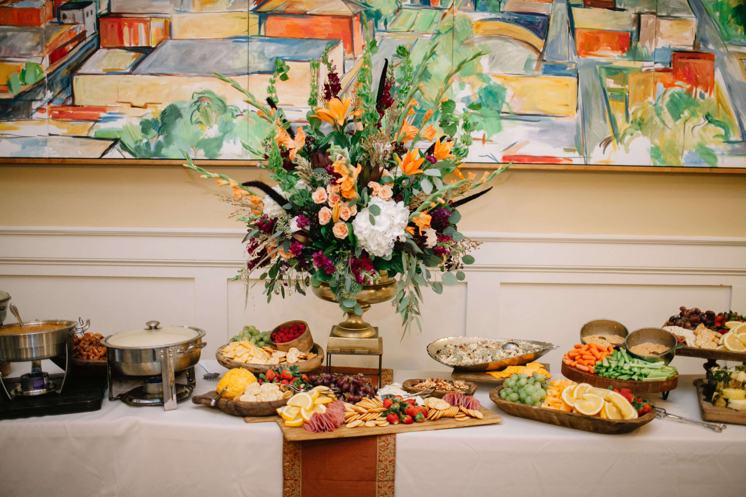 A buffet table is set with assorted cheeses, fruits, vegetables, crackers, and hot dishes. A large floral centerpiece with orange, pink, and burgundy flowers stands in the middle.