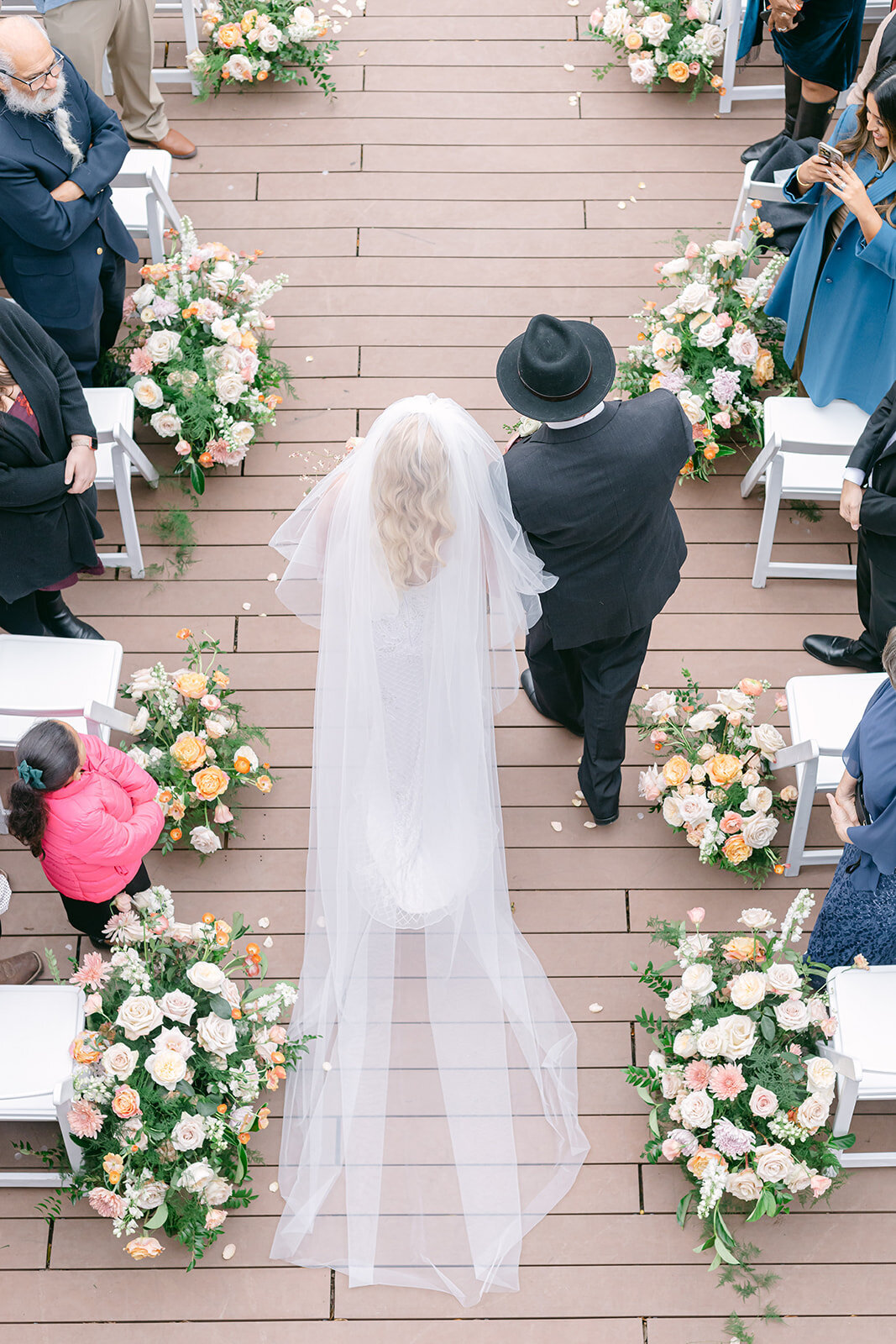 Bride walking down a wooden floral-lined aisle with her father at a romantic outdoor Austin wedding ceremony, guests looking on as soft natural light fills the space