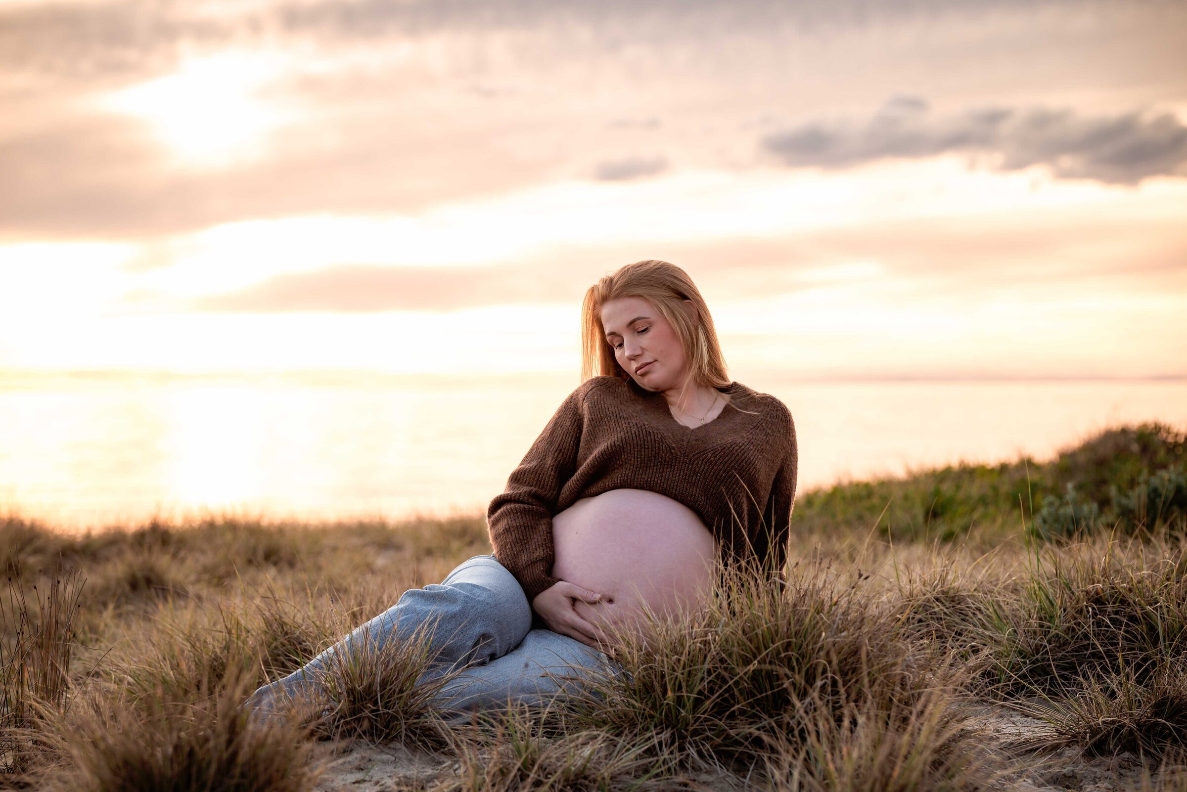 Pregnant mother in blue jeans and a brown jumper sitting on the dunes at the beach with the sun setting behind by Melbourne Family Photographer Kate Cashin