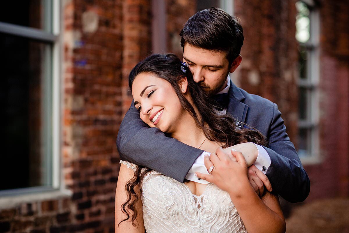 Groom hugging his wife and snuggling with her outside of historic Cannery ONE brick walls