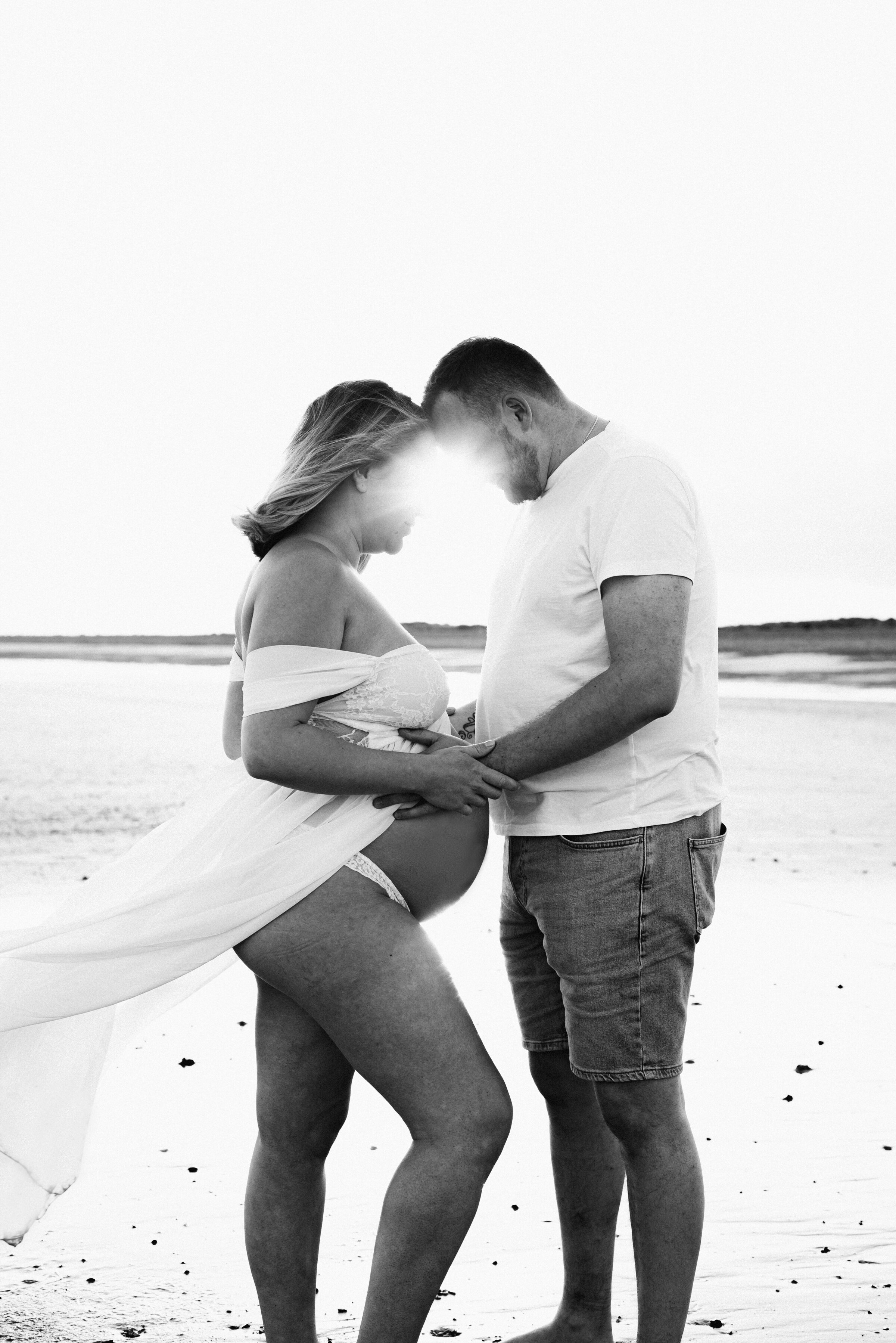 Mother and father holding pregnant mothers belly looking down on the beach in Billingshurst at maternity photoshoot
