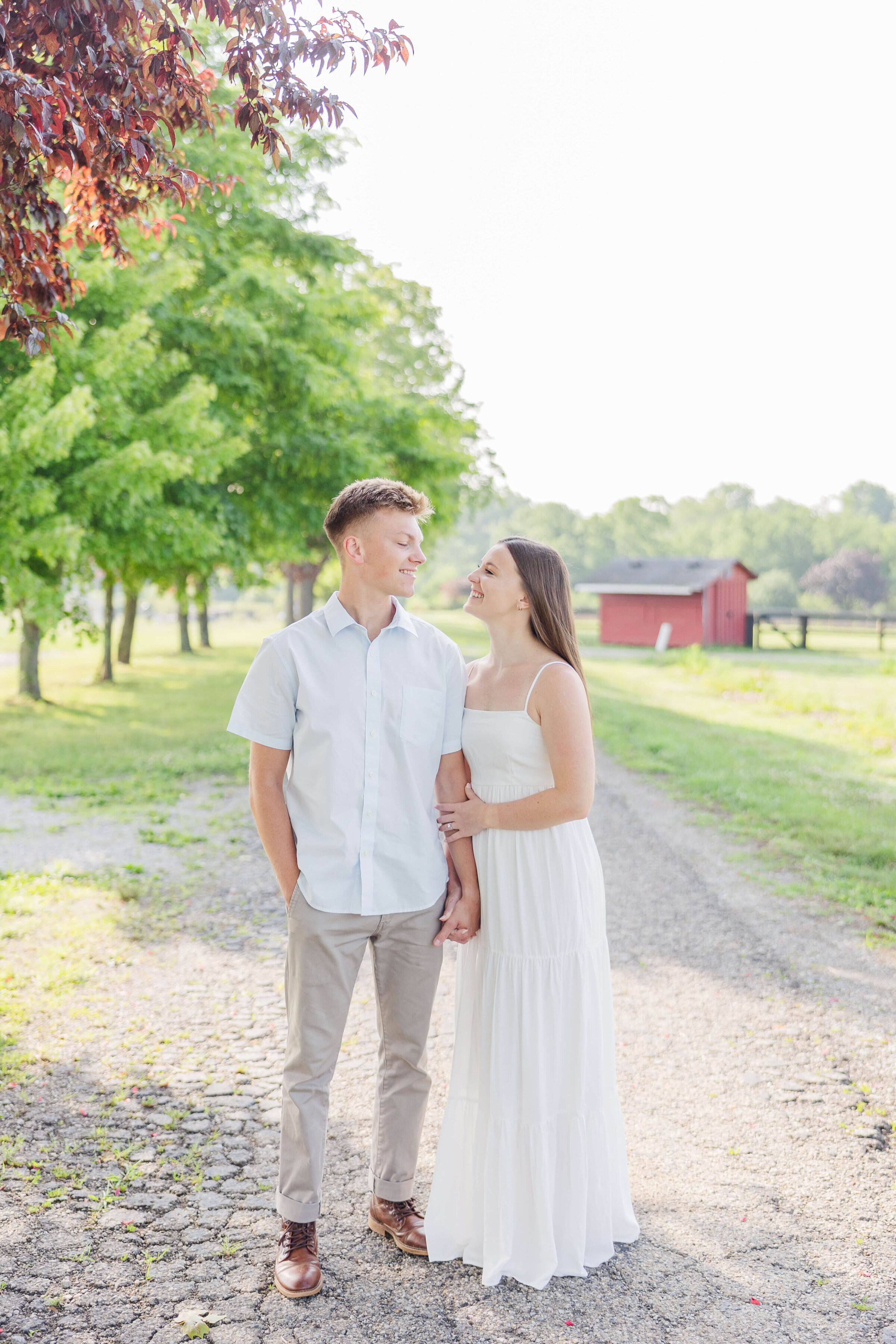 A man in khakis and a light blue shirt holds hands with his fiancee who is wearing a white dress. They are walking down an orchard path as the sun shines brightly in back of them.