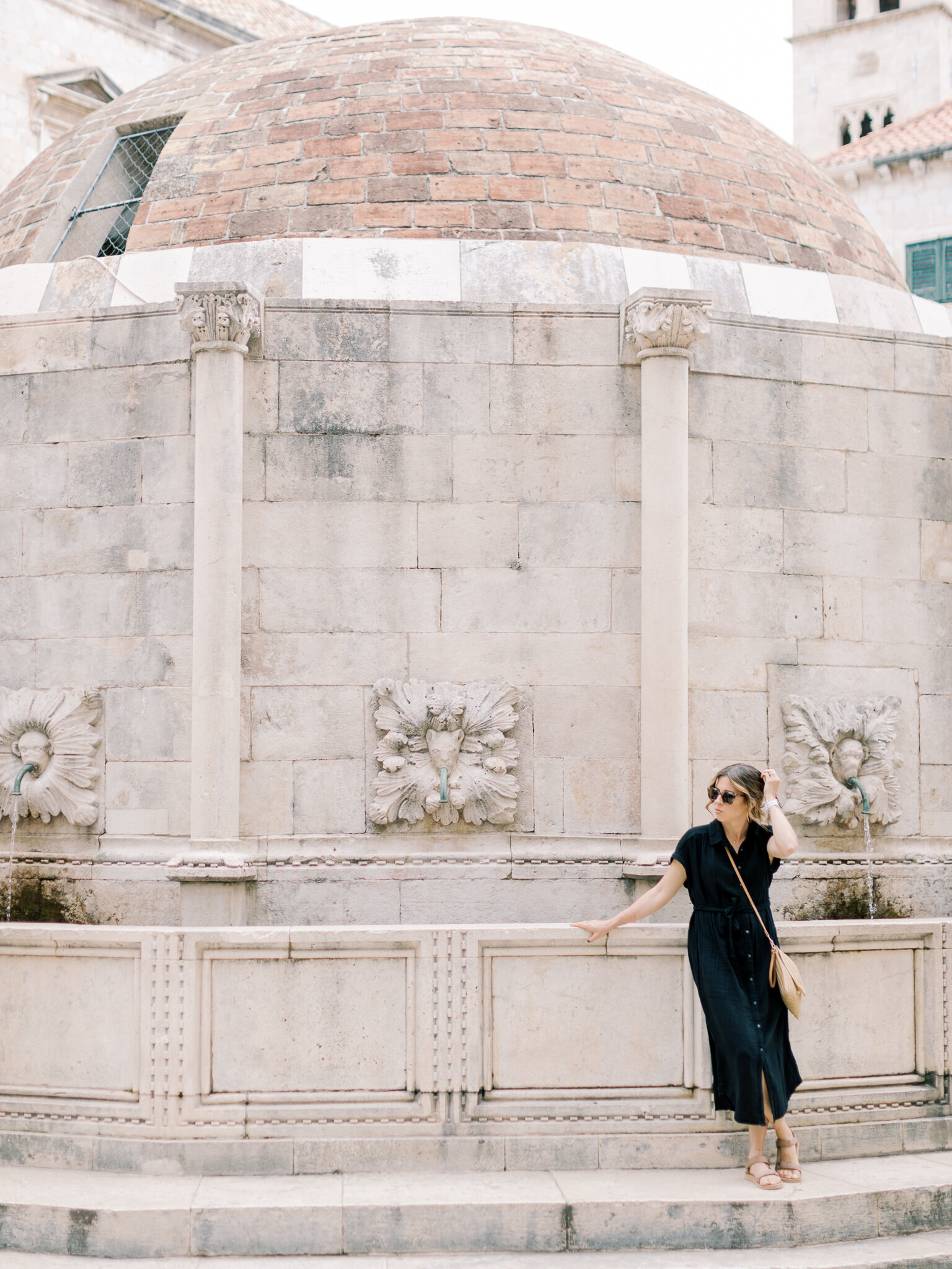 Little Rock photographer Bailey Feeler in black dress and sunglasses stands next to Croatian fountain