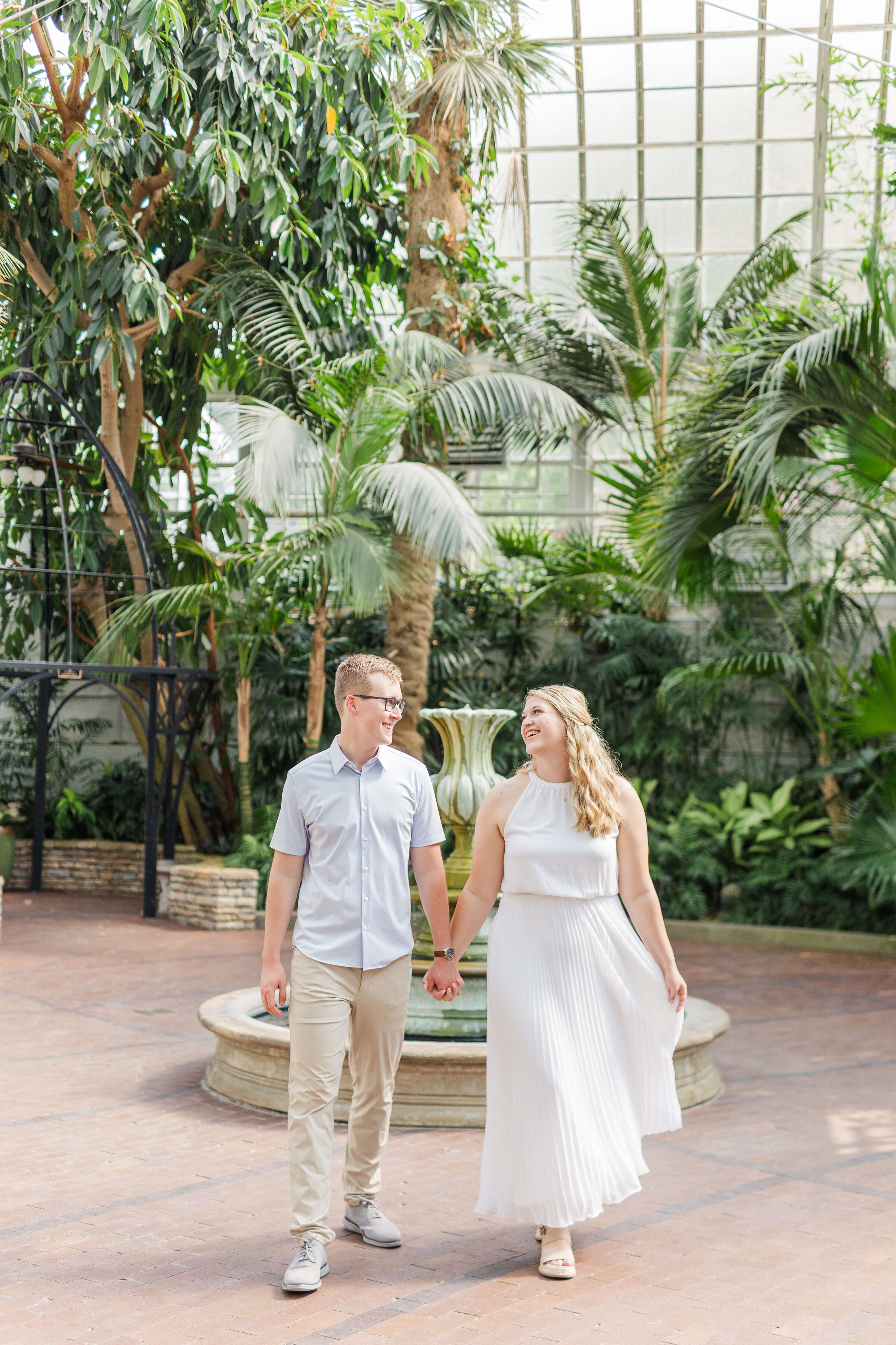 A man and woman hold hands as they walk in a conservatory. There are lots of green plants around. She is wearing a white dress and he is wearing khaki and a blue shirt.