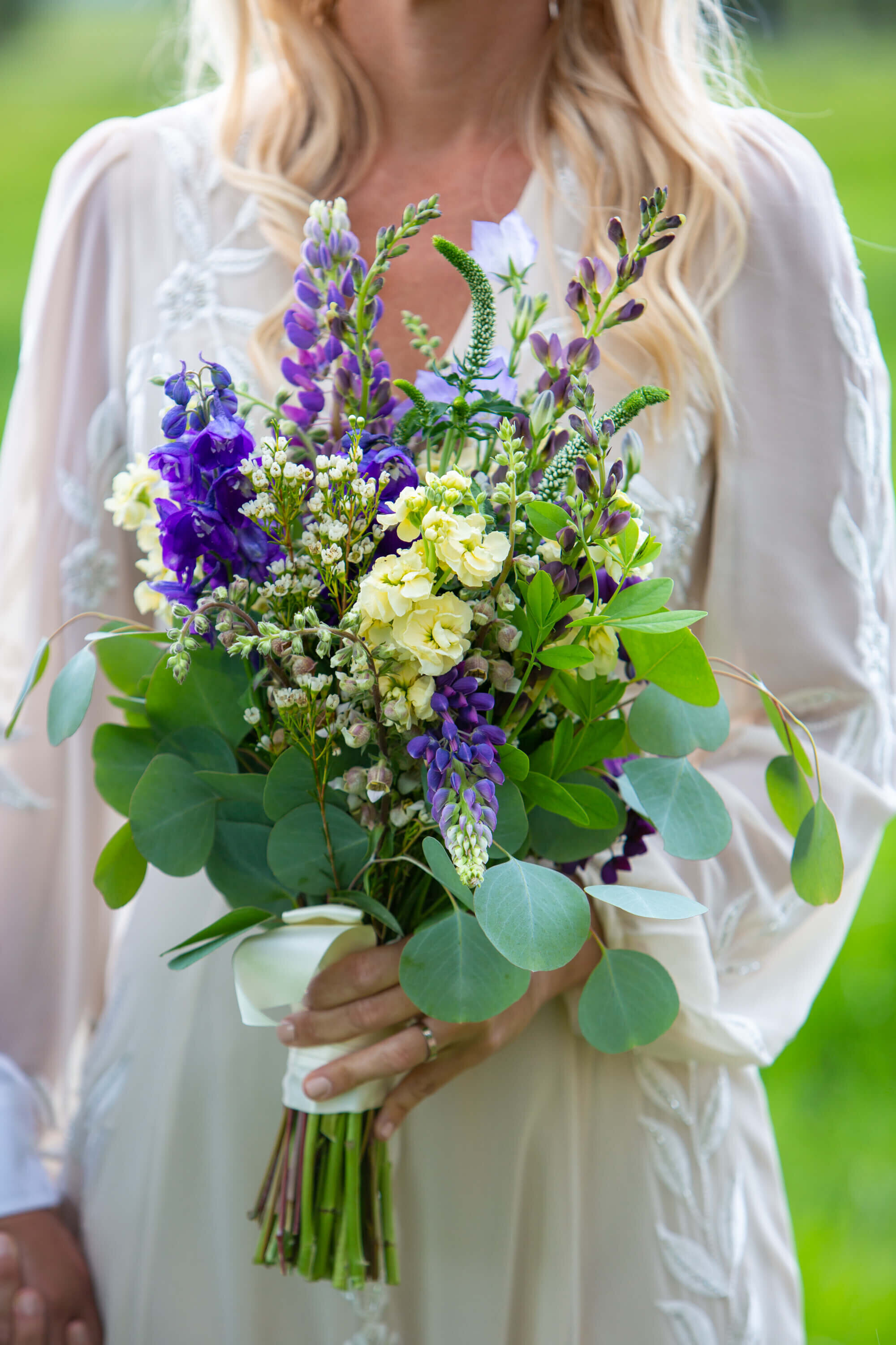 A bride holds a bouquet of wildflowers in shades of purple, yellow, and white, accented by lush greenery, wrapped in a white ribbon. Her flowing wedding gown features delicate lace sleeves. The outdoor wedding setting in Jackson Hole, Wyoming, provides a soft, natural backdrop.