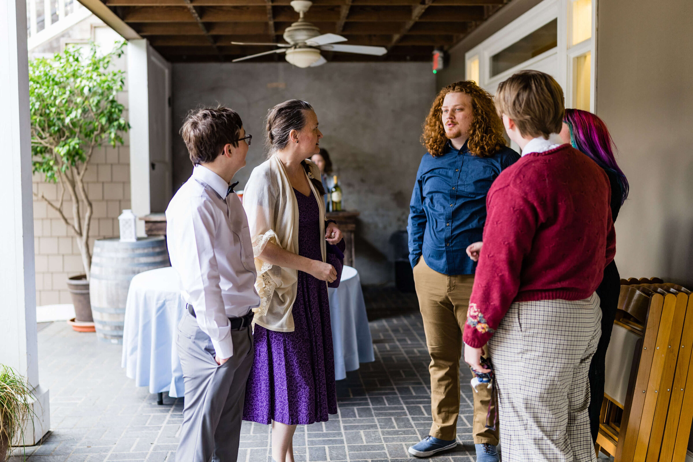Five people stand in a casual group indoors, engaged in conversation. One person wears a purple dress, others wear shirts, pants, and sweaters. Tables, folding chairs, and a barrel are in the background.