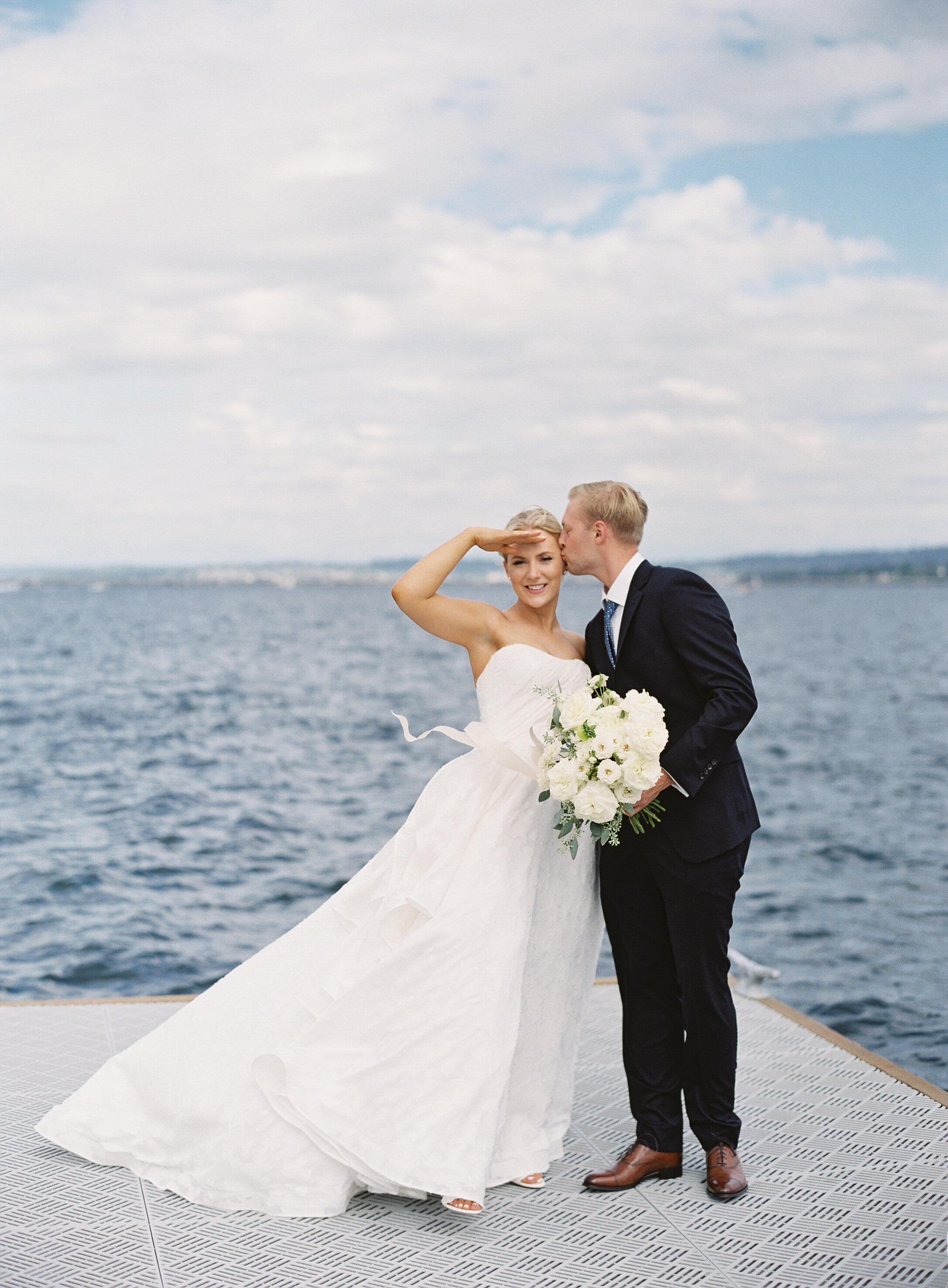 bride holds white floral boquet while sheilding sun from face on dock overlooking blue water while groom kisses her cheek