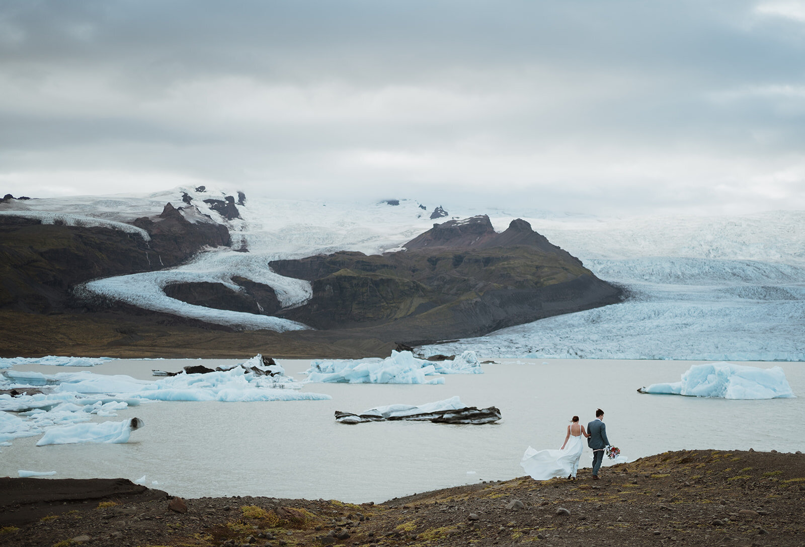 the bride and groom are walking along a ridge that leads to a basin filled with glaciers and mountains. they are far away and the bride's dress is blowing in the wind. the groom is holding her flowers as they walk side by side.