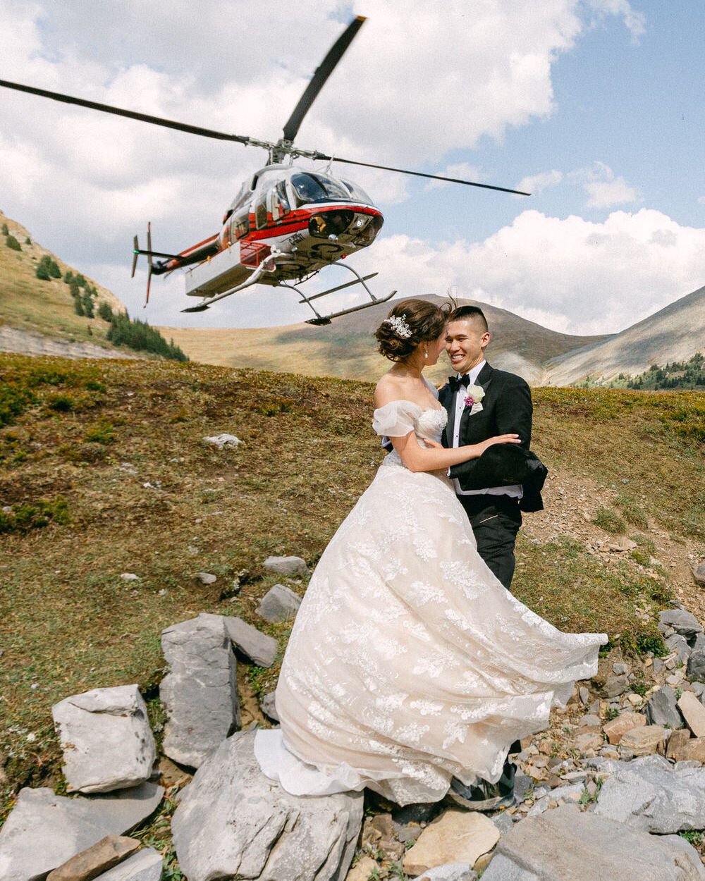 An epic adventure elopement moment as the helicopter lifts off behind the bride and groom in Banff National Park. With laughter and joy, they hold each other, embracing the excitement of their intimate mountaintop ceremony. The roaring helicopter overhead adds a dramatic and unforgettable touch to their once-in-a-lifetime experience in the Canadian Rockies.