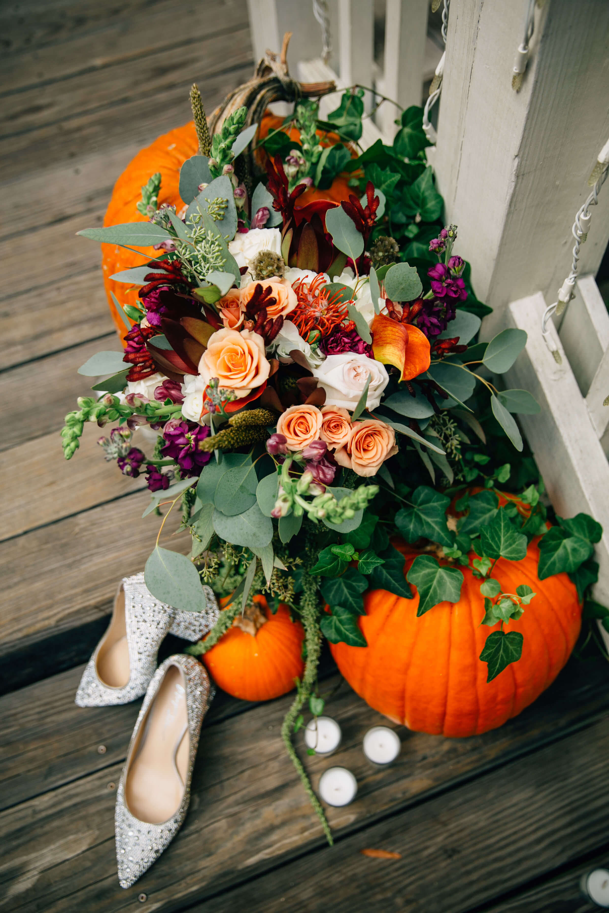 A colorful bouquet of flowers and greenery rests on two pumpkins beside white candles and sparkly high-heeled shoes, all arranged on a wooden floor near a white fence.