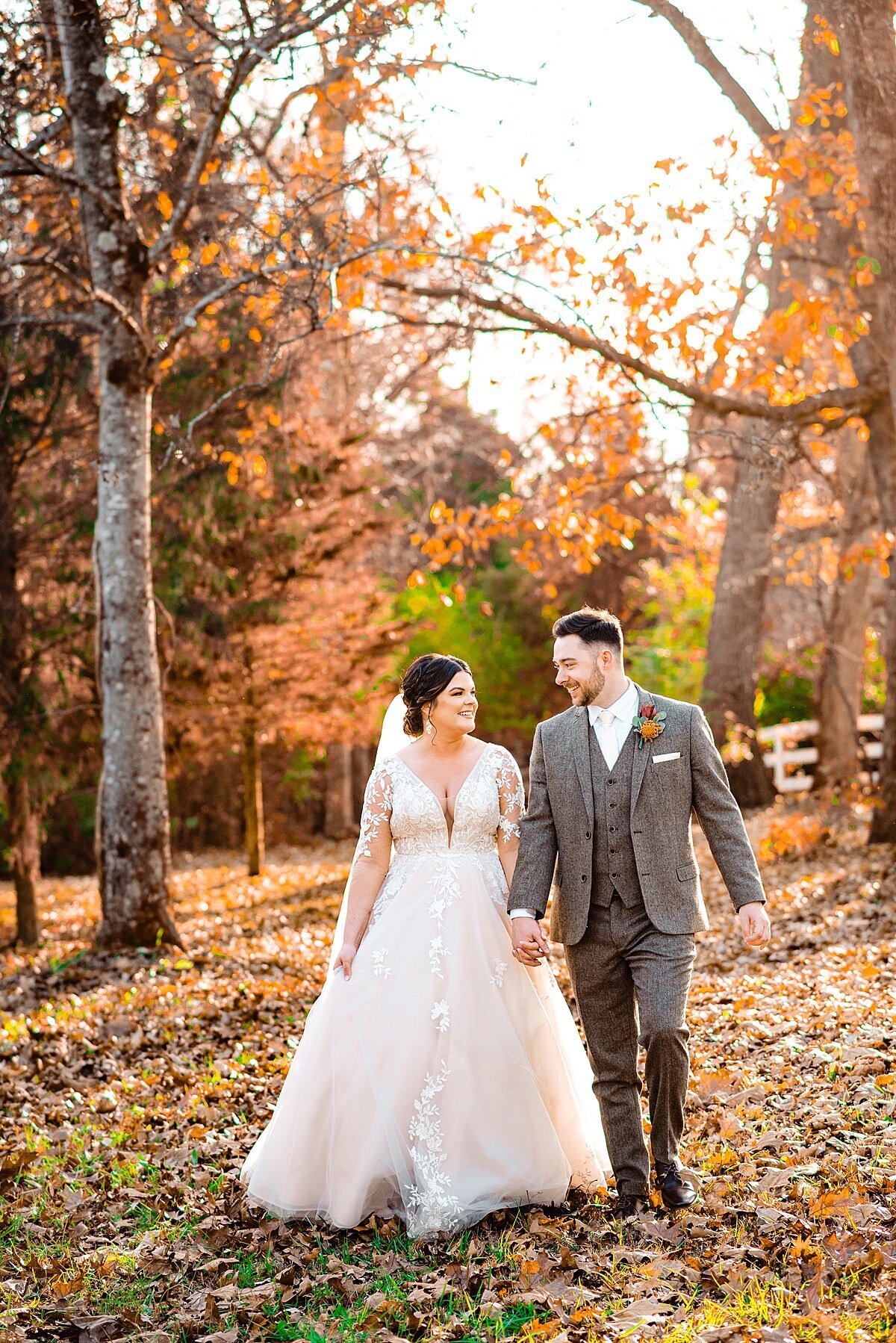 Bride wearing long sleeve lace wedding dress walking hand in hand with groom who is wearing a grey tweed suit strolling through leaves outside of Copper Ridge event center