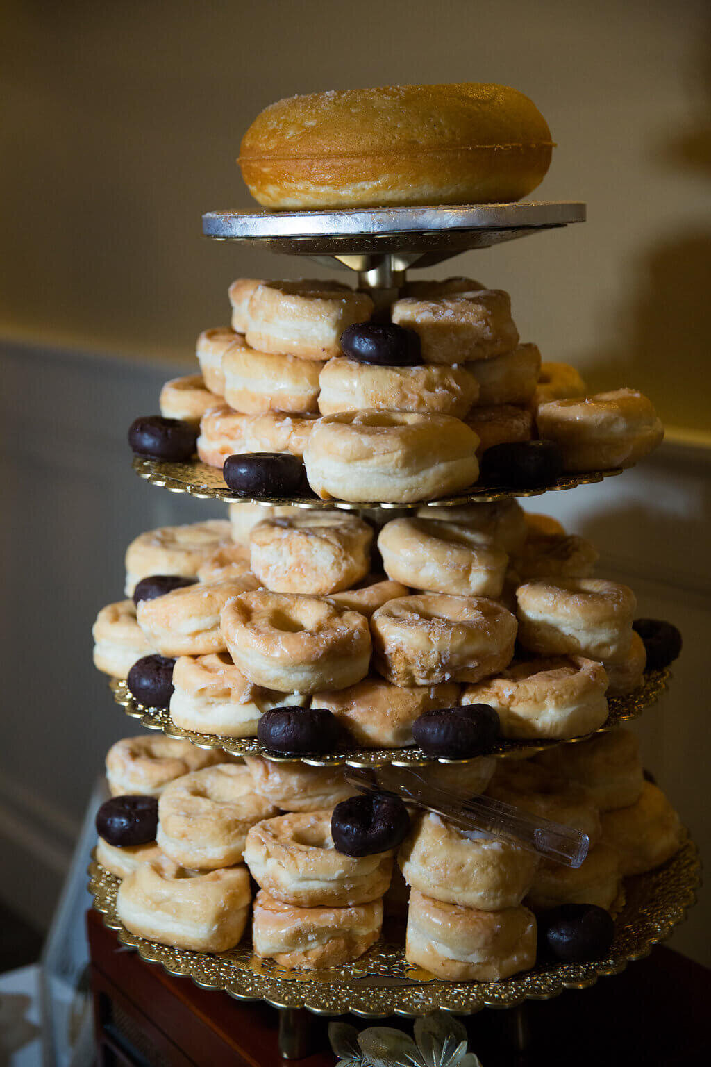 A three-tiered gold stand filled with stacks of glazed doughnuts and a few chocolate doughnut holes, topped with a single large glazed doughnut on the top tier.