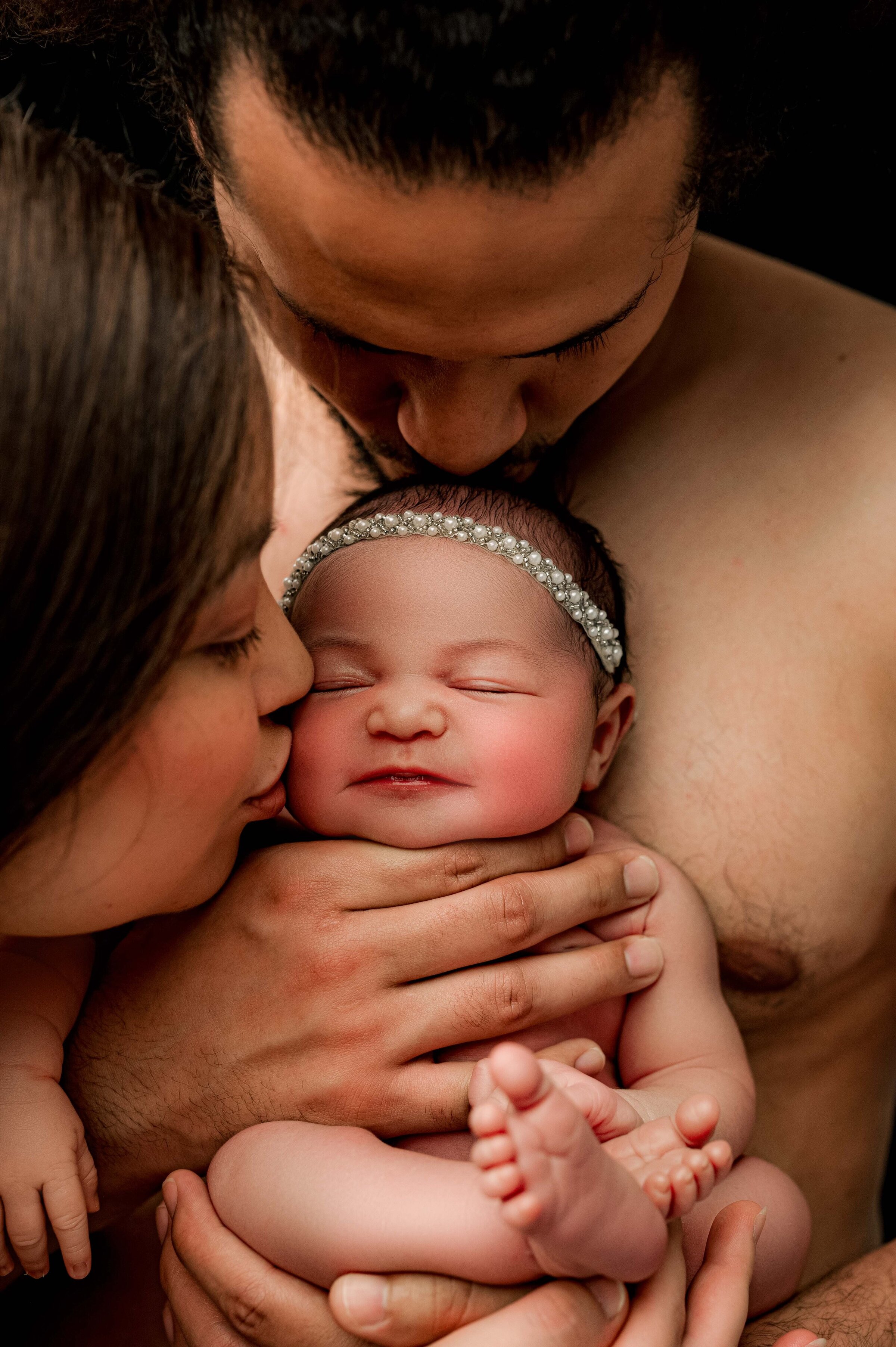 A couple lovingly holds their newborn baby, captured beautifully by a North Carolina newborn photographer. The baby, adorned with a silver headband, smiles gently in their arms. The woman kisses the babys cheek while the man leans in close, creating a warm and intimate scene.