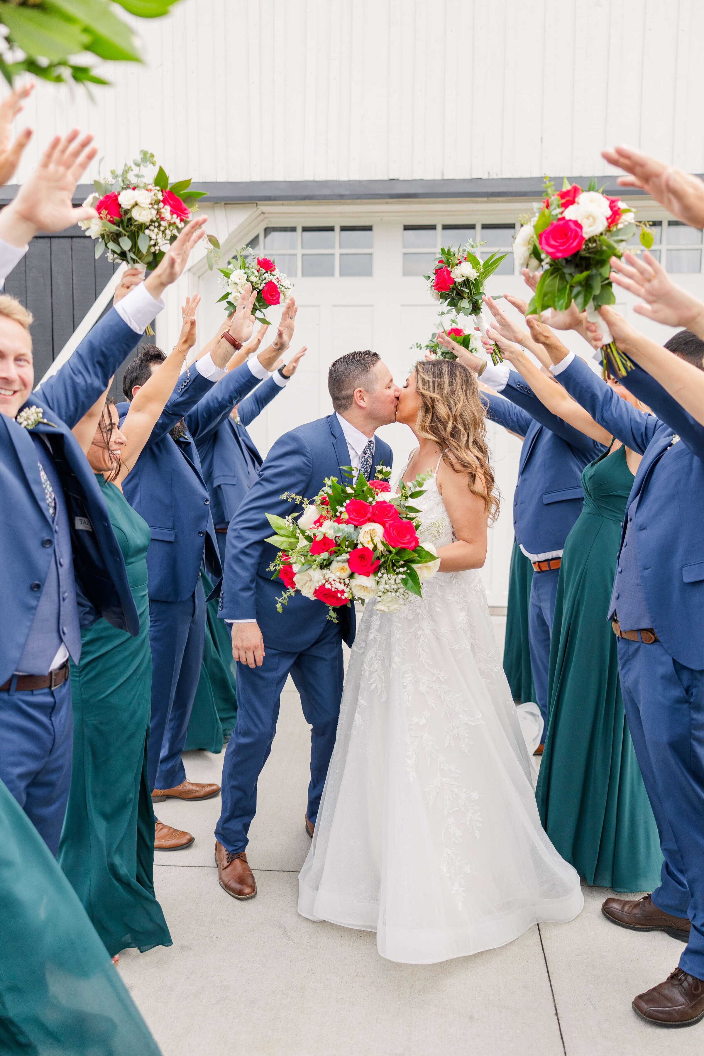 A bride and groom kiss as their bridal party cheers for them. The groomsmen are in blue suits and the bridesmaids are in teal dresses.