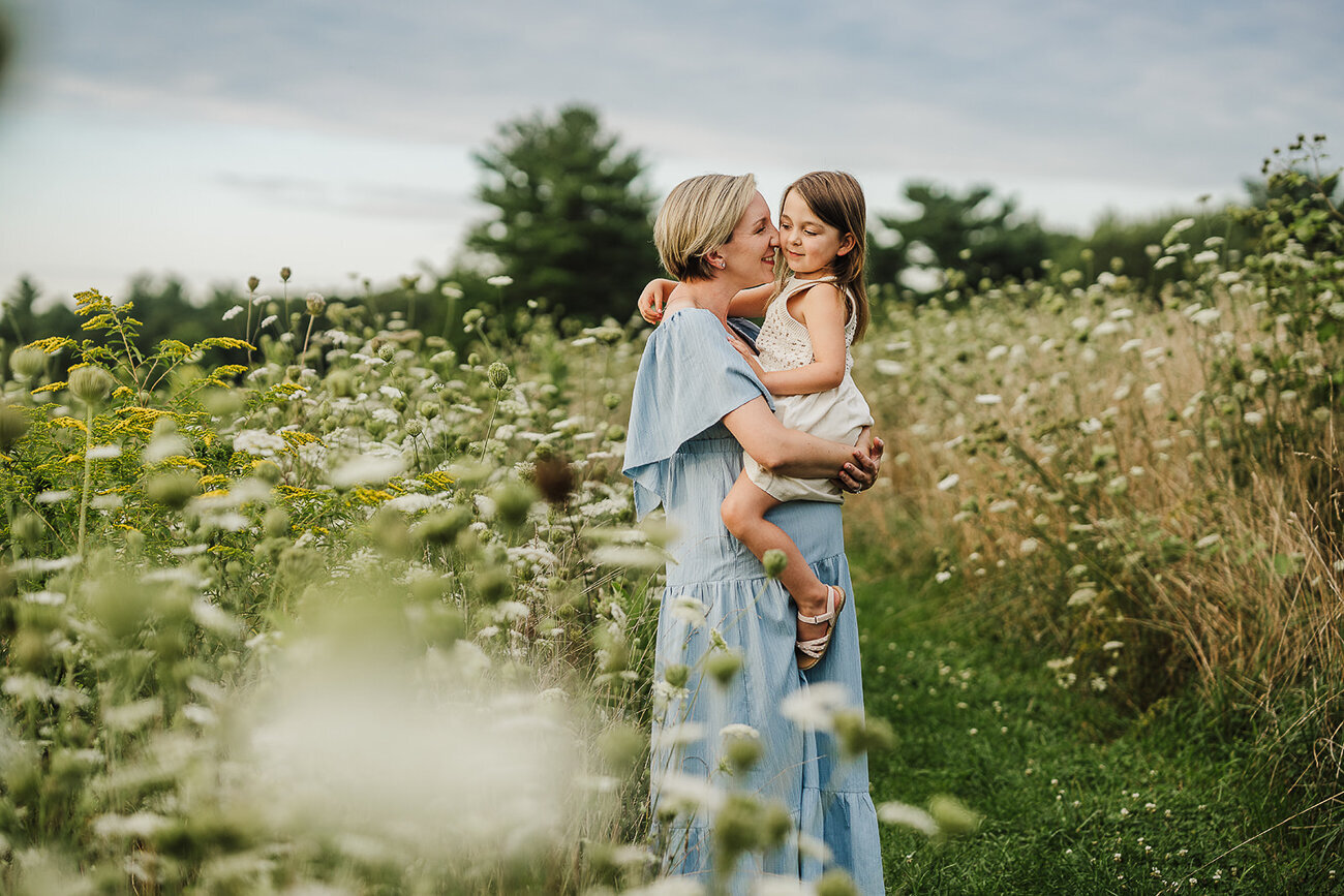mother snuggles daughter in field of queen annes lace during maternity photoshoot in boston