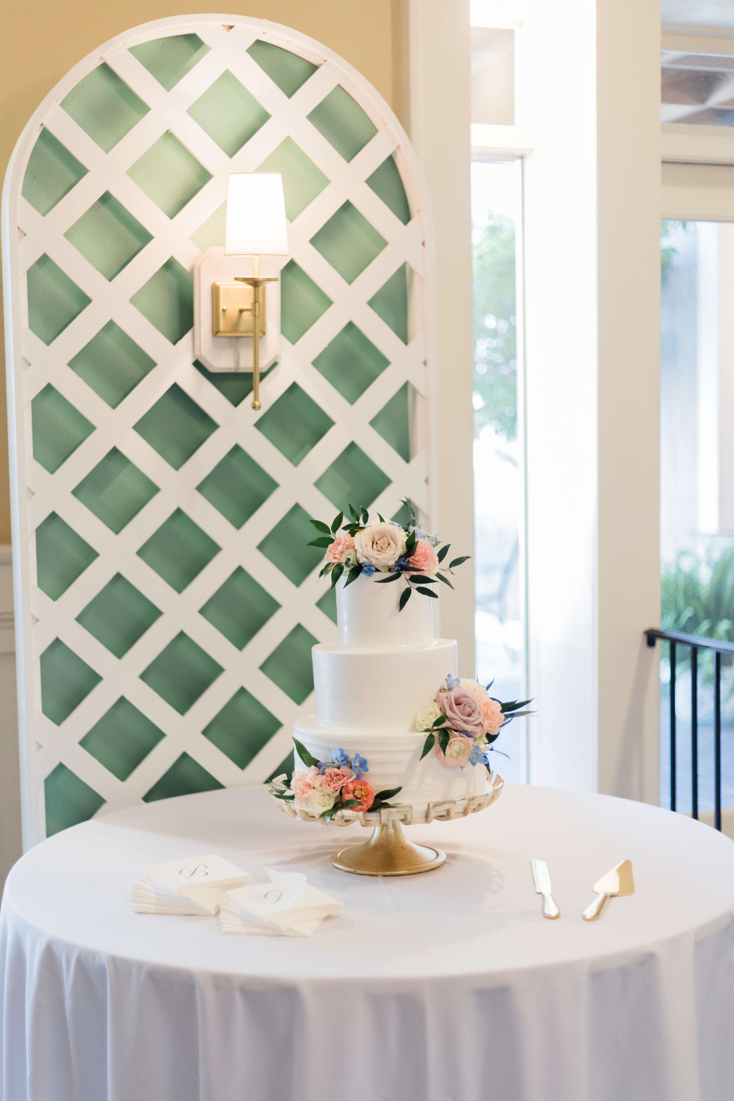 A white tiered wedding cake decorated with pastel flowers sits on a round table with a white tablecloth. Cake-cutting utensils and napkins are nearby. Behind is a green lattice wall with a white sconce.