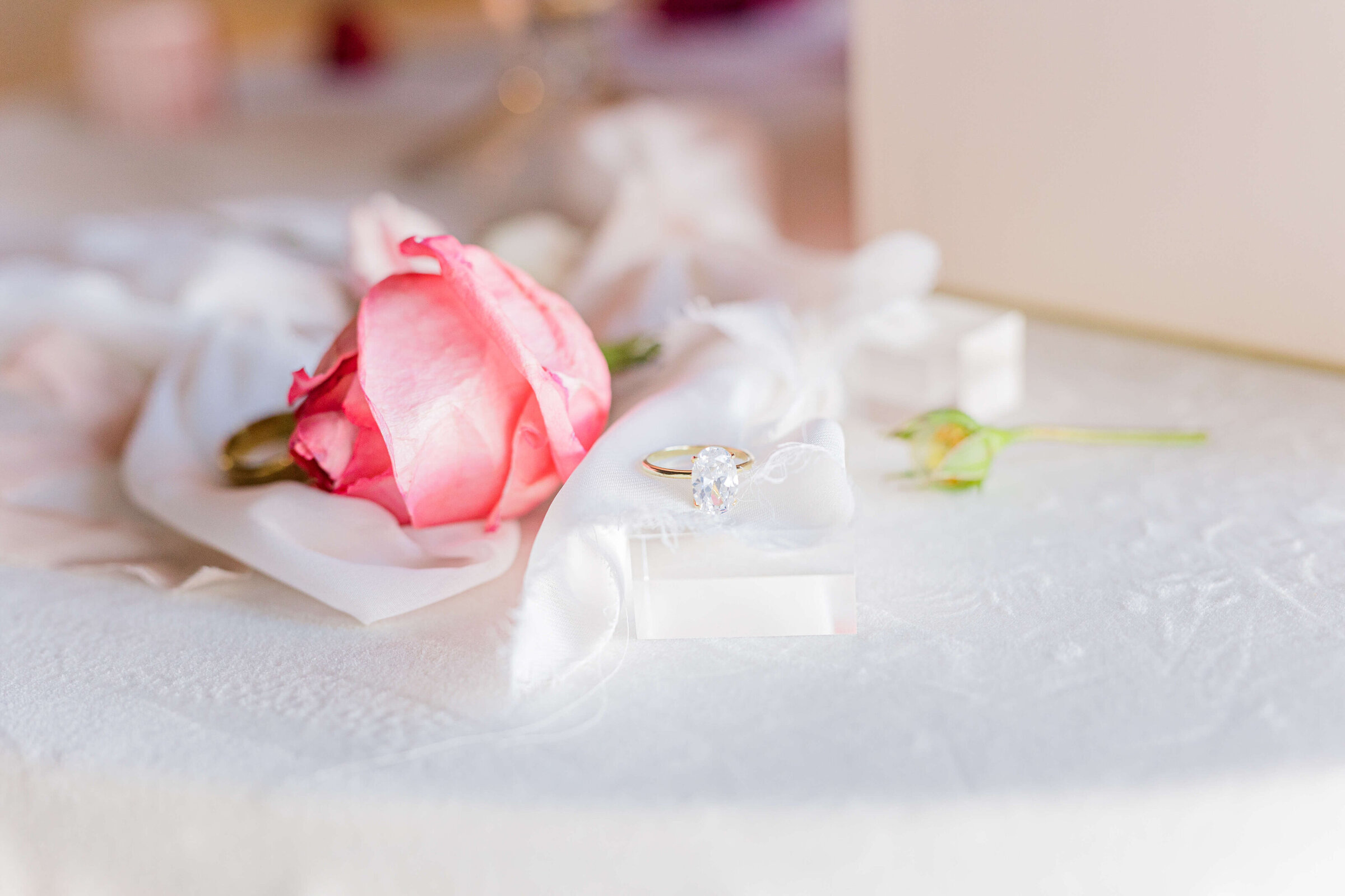 A pink flower sits on a table with a white tablecloth and a diamond ring
