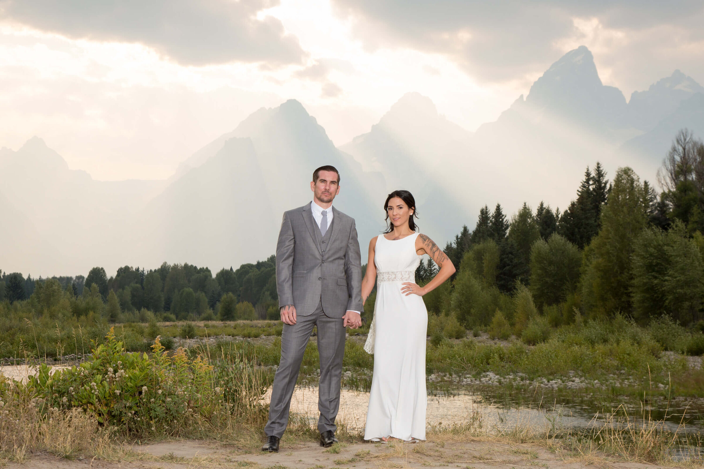 Grand Teton wedding portrait – A bride in a sleek white gown and a groom in a gray suit pose in front of the Grand Teton mountains. Sunbeams break through the clouds, creating a dramatic wedding backdrop.