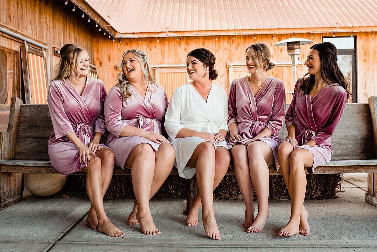 Bride with bridesmaids wearing robes sitting on bench outside