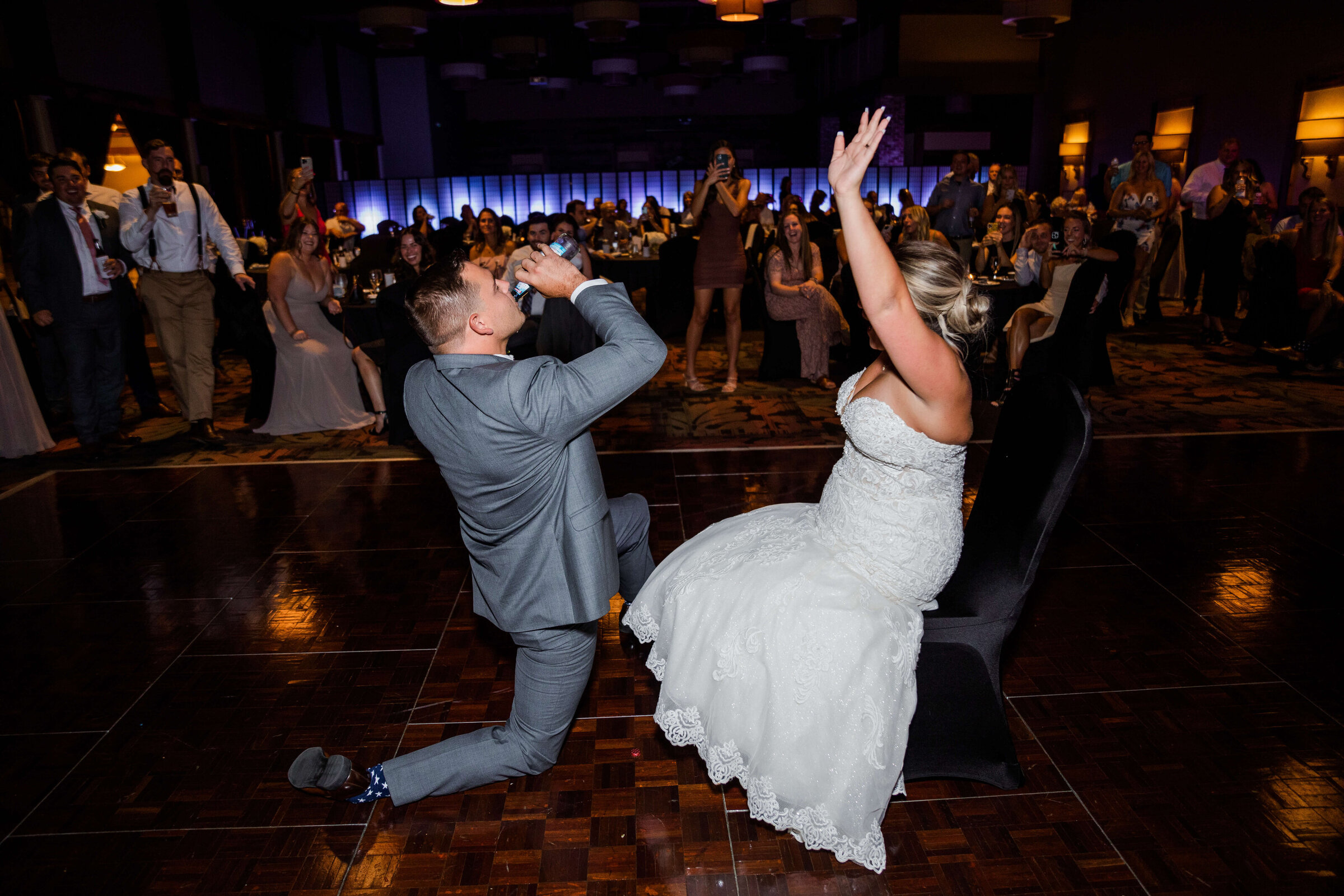 A groom is drinking a smirnoff ice on the dance floor while on one knee while his bride sits in a chair cheering him on