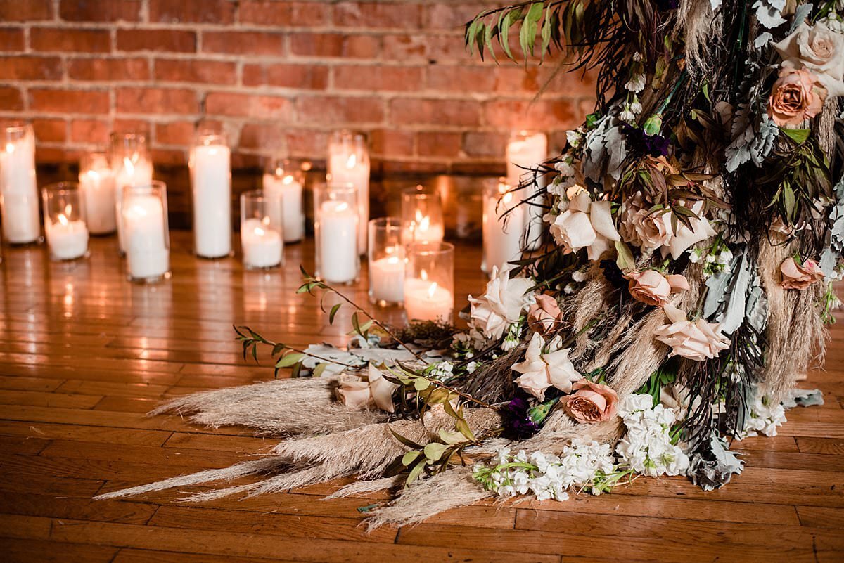 Boho flower arrangement on the wooden floor with pampas grass and roses, candles of different heights surrounding it and bricks in the background