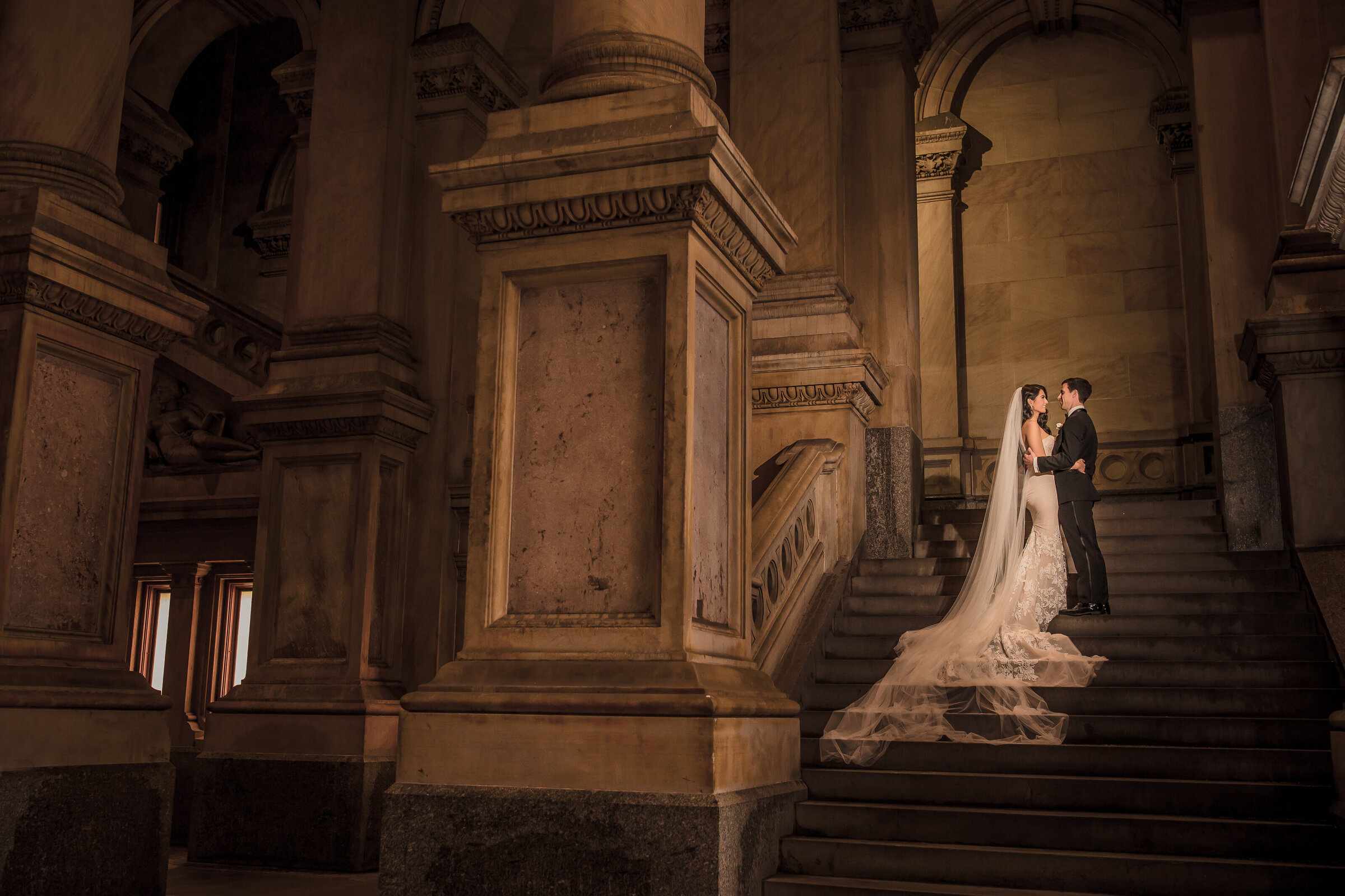 Bride with long cathedral veil and the groom embrace on the grand staircase at Philadelphia City Hall in dramatic light