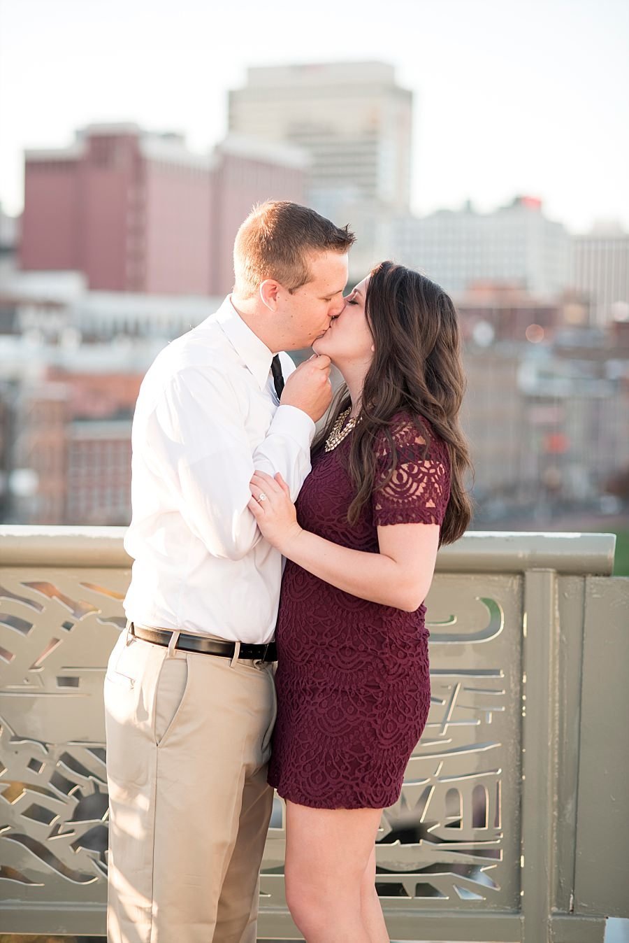 Girl wearing a burgundy lace dress kissing her fiance on the bridge in downtown Nashville with the city behind them