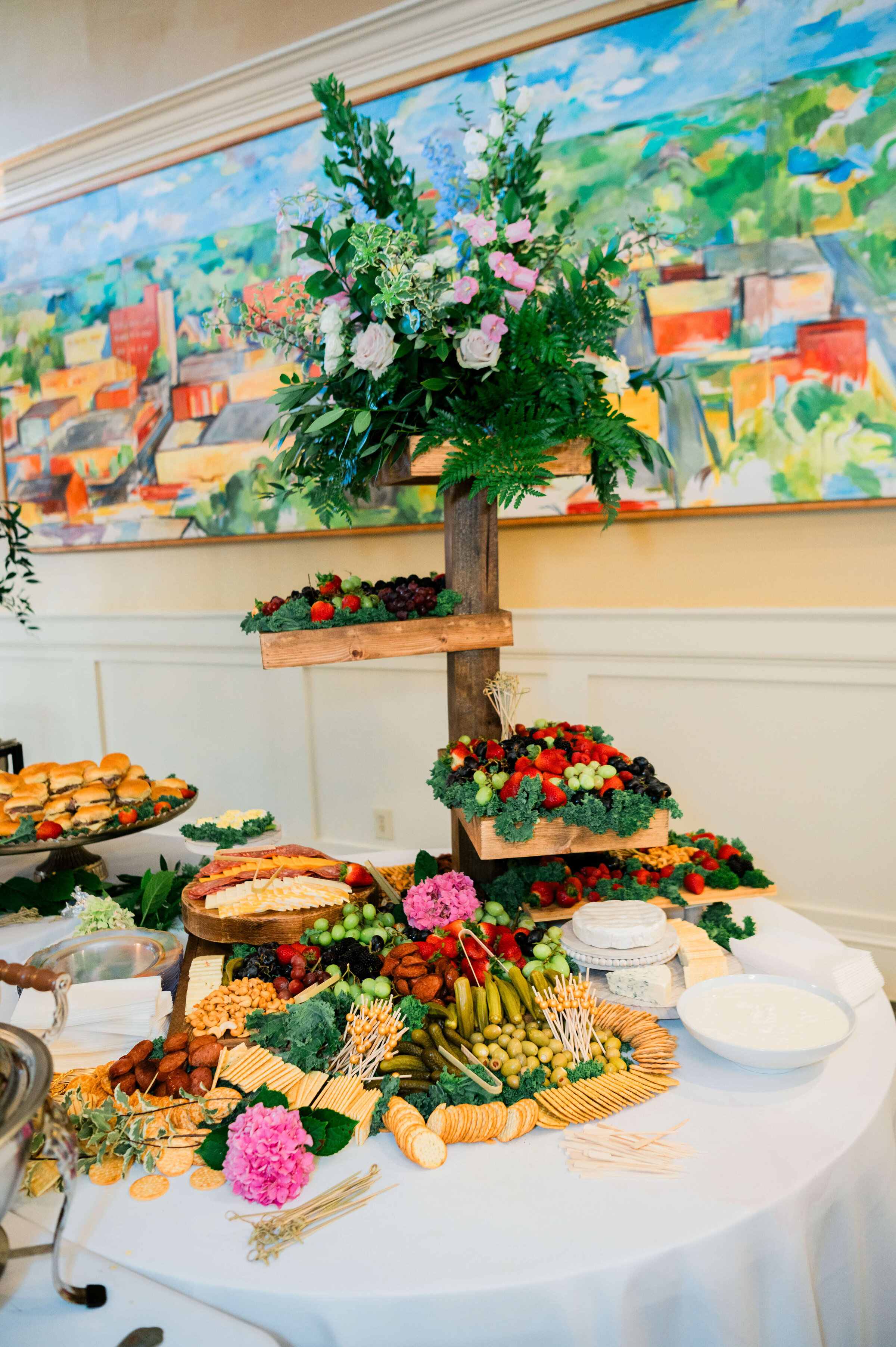 An elegant display of assorted cheeses, crackers, fruits, vegetables, and charcuterie, topped with a vibrant floral arrangement and two wooden shelves holding more fruit.