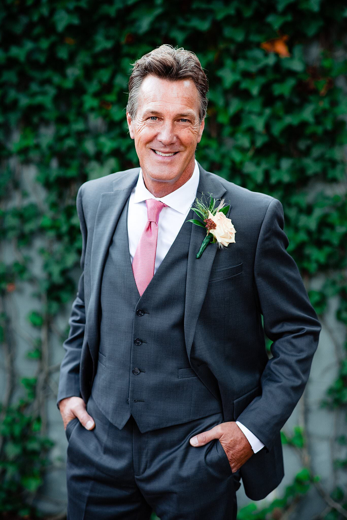 Groom wearing a grey suit and coral tie standing in front of an ivy covered wall