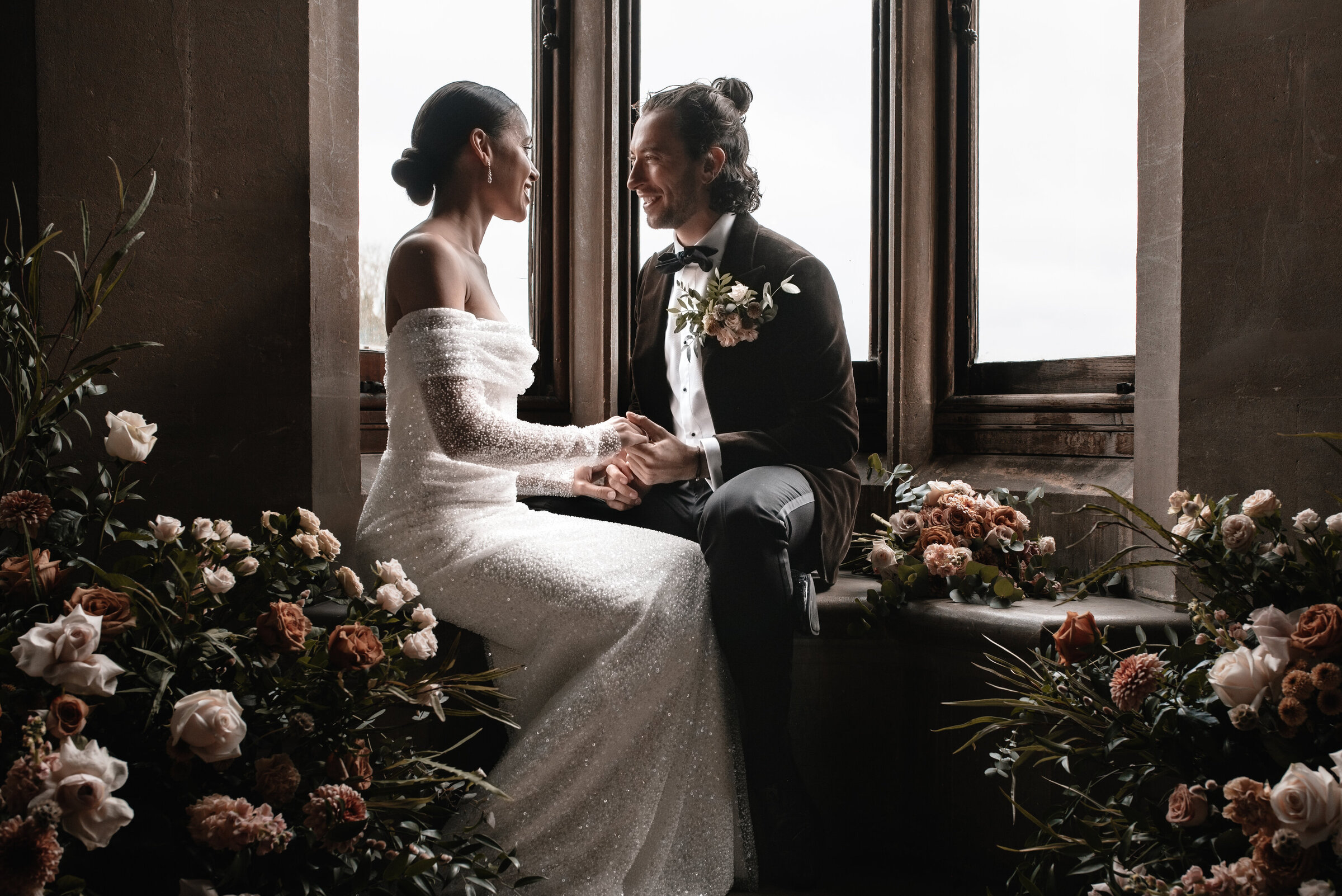 Bride and Groom in wedding attire talking in the windowsill surrounded by floral arrangements