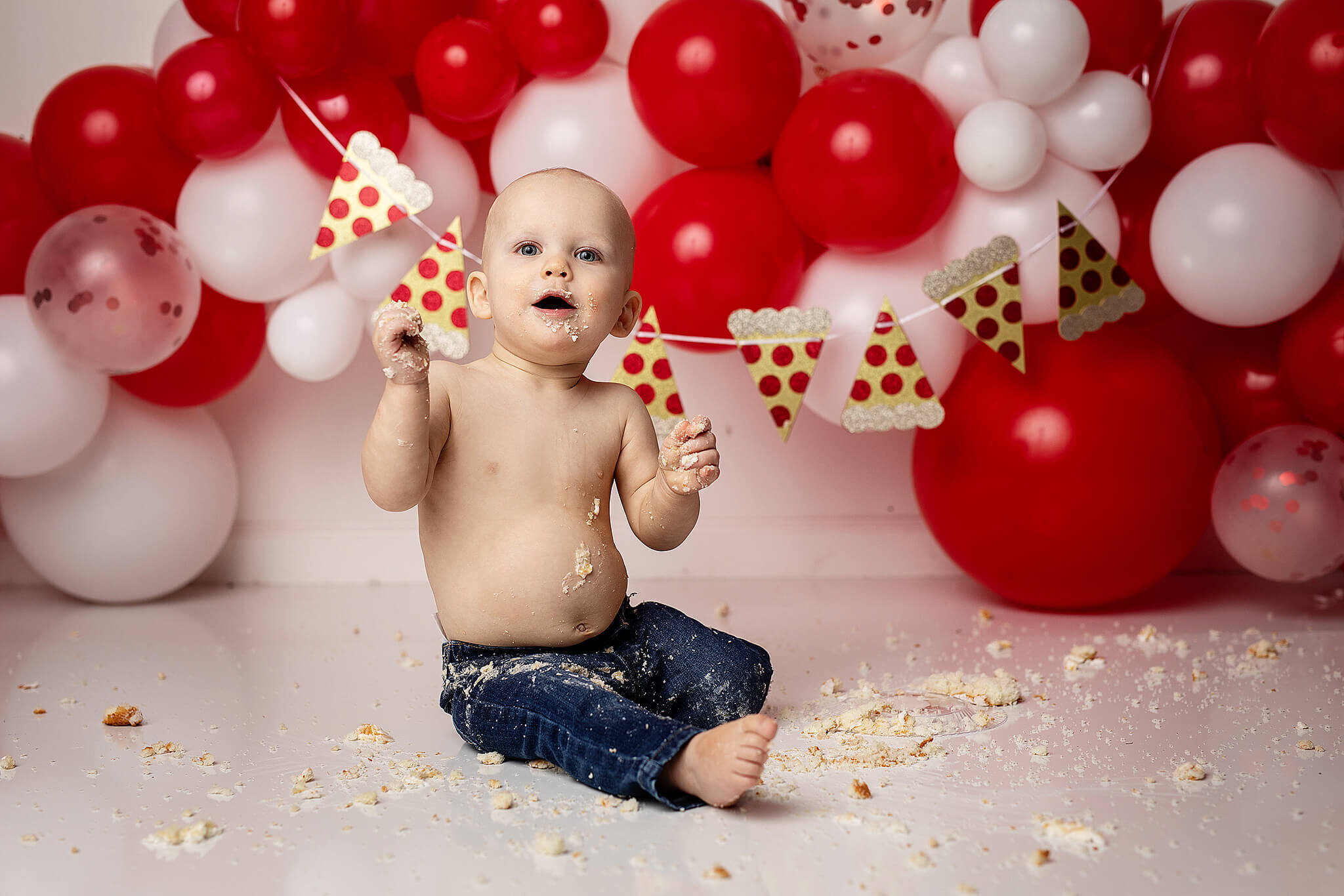 Little boy with red and white balloons and a pizza banner behind him, while he is smashing a cake to pieces. 