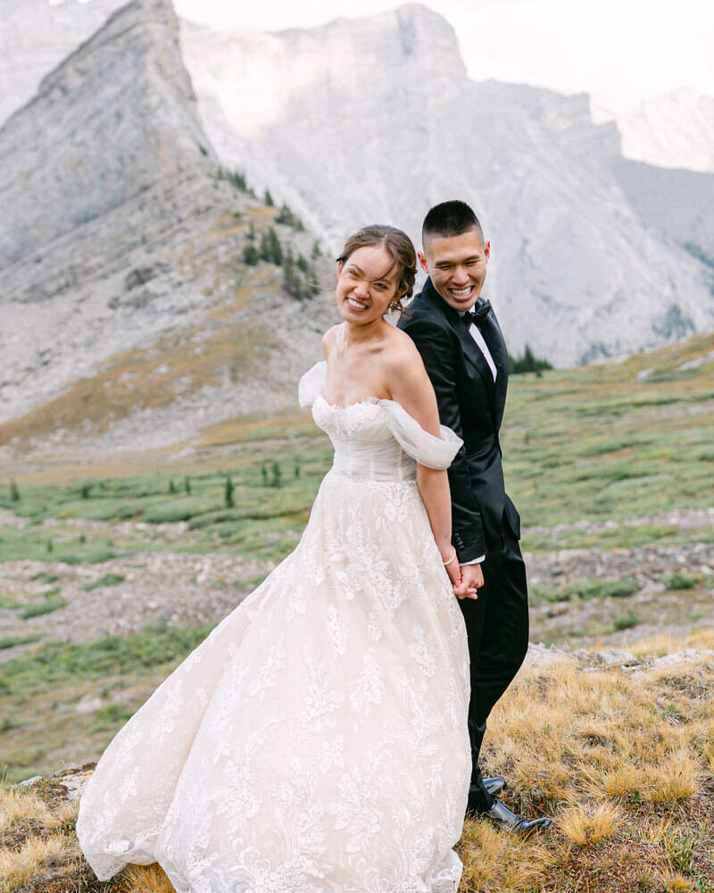 An adventure-filled alonement photograph capturing the bride and groom back-to-back, laughing joyfully amidst the towering mountains of their heli-hiking elopement in Banff National Park. With breathtaking alpine scenery surrounding them, this moment embodies the excitement, connection, and pure joy of their unforgettable mountain wedding experience.