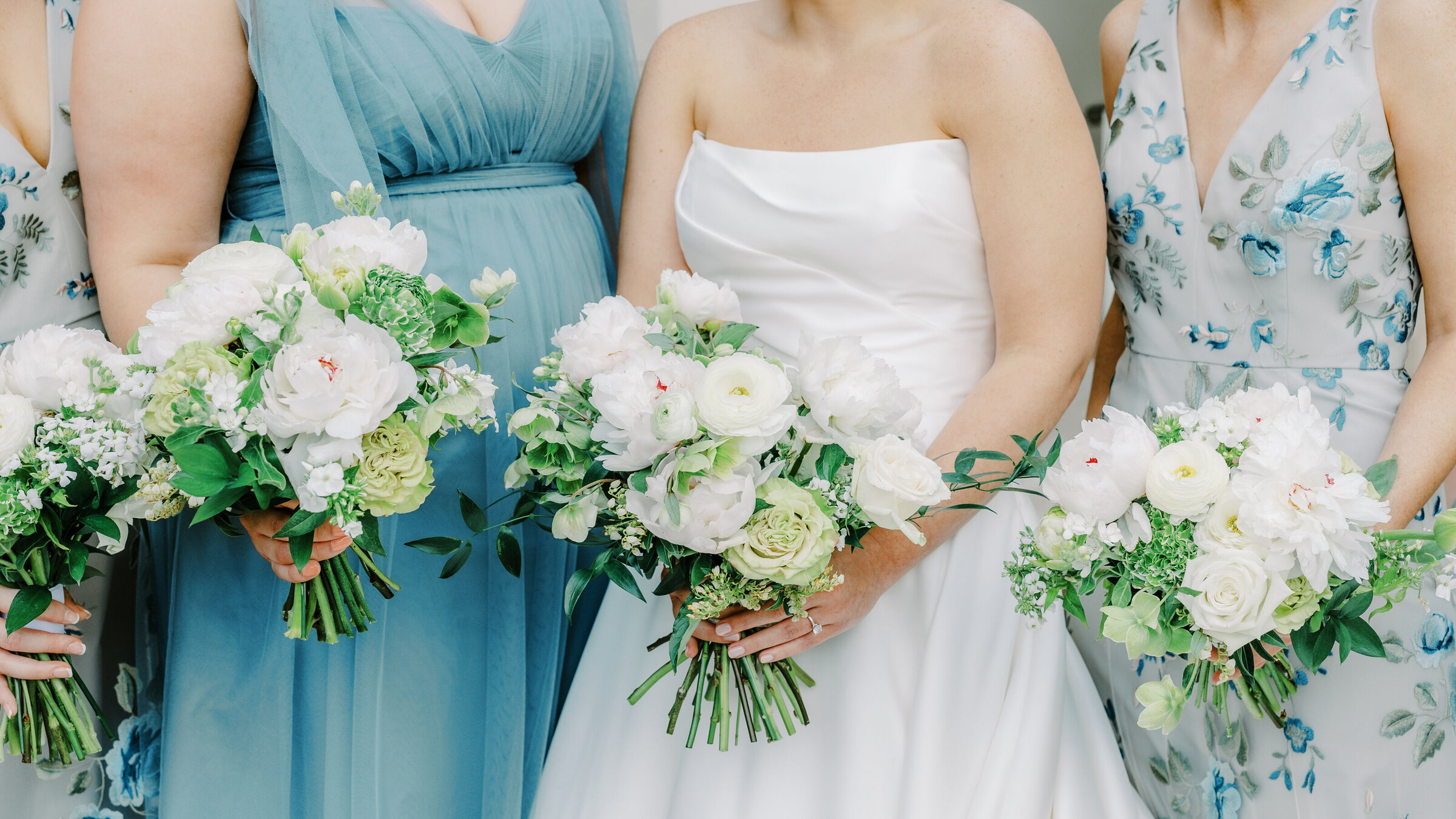 Bridesmaids in blue and green floral dresses and bride in white strapless dress holding bouquets with white ranunculus