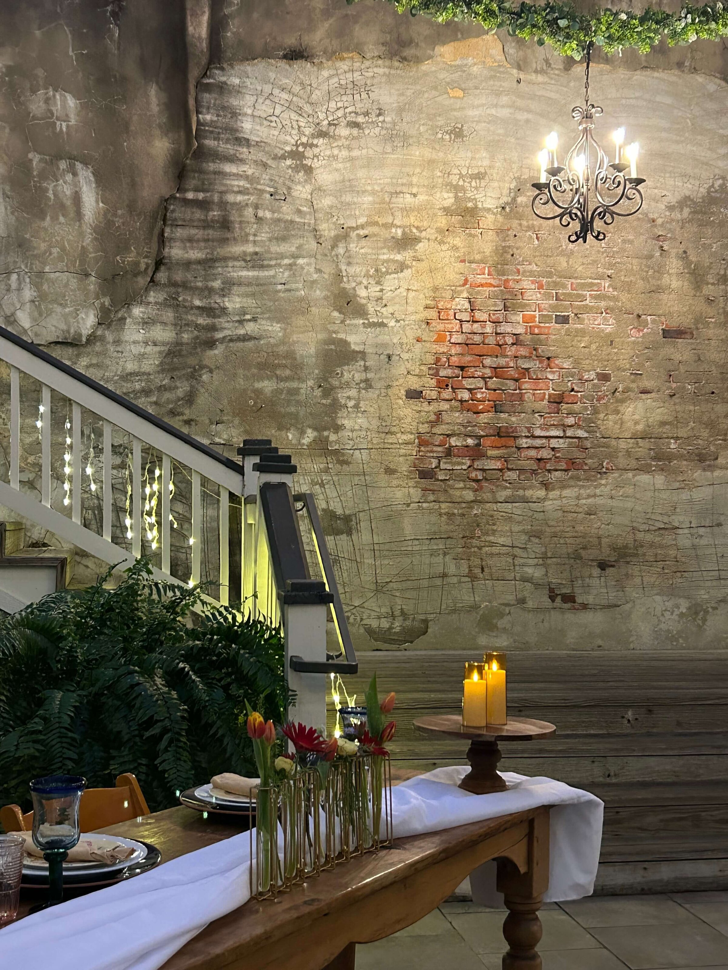 A romantic outdoor scene with a brick and plaster wall, a chandelier, a wooden table set with flowers, candles, and tableware, green plants, and a white staircase decorated with lights.