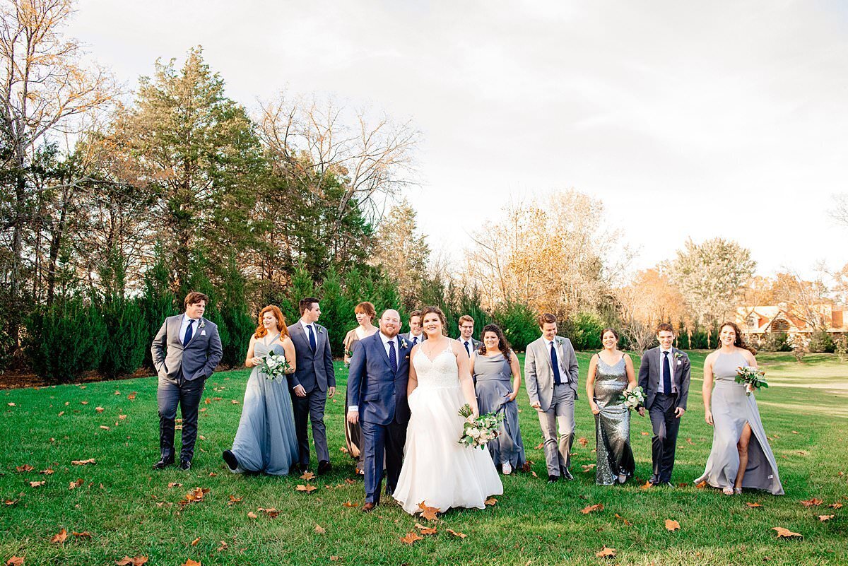 Wedding party wearing shades of blue and grey  walking together in the field at Sycamore Farms, fall tree beginning to drop leaves