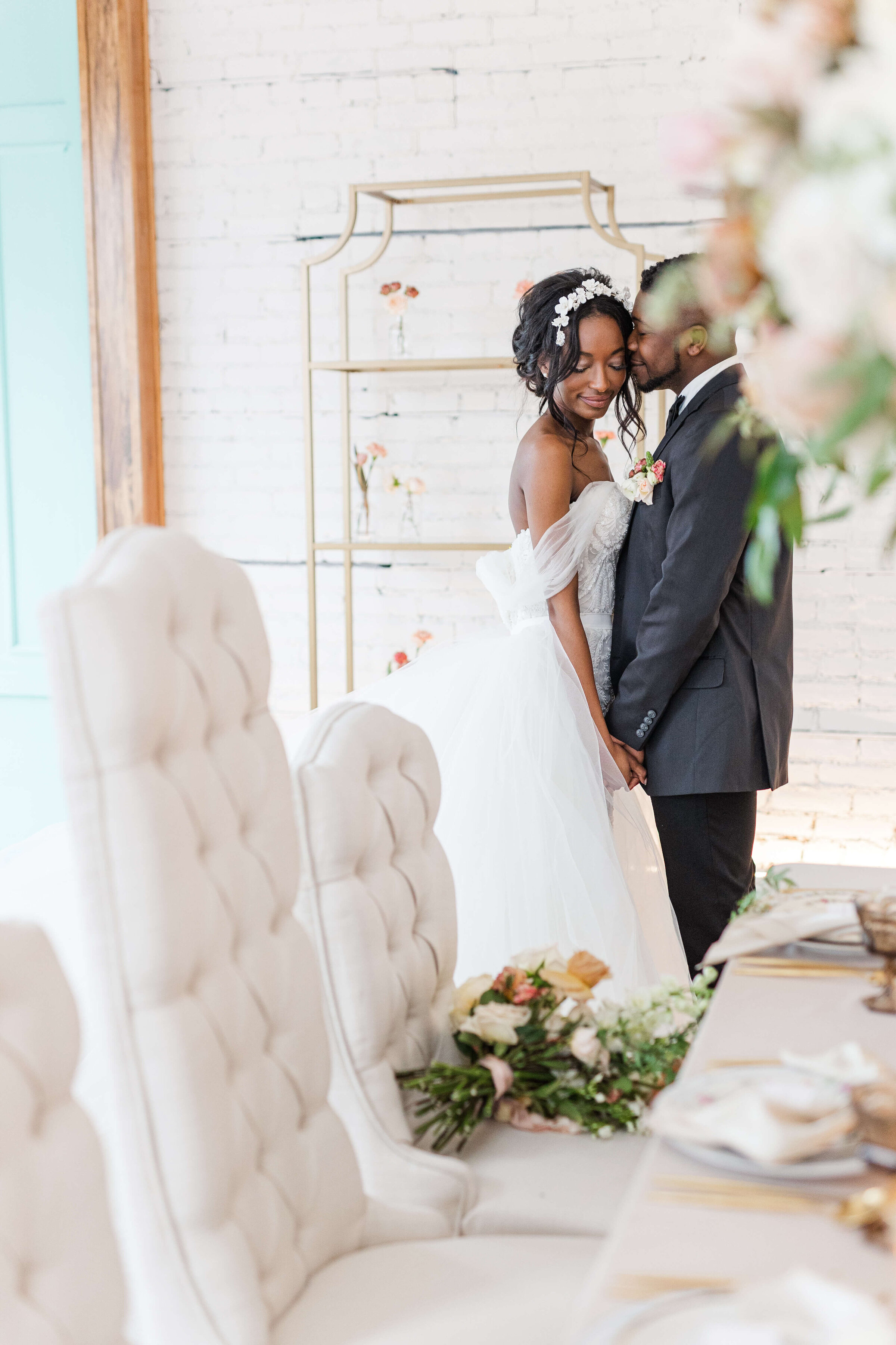 A bride stands in front of her groom as he kisses her check and she looks down. They are standing inside by their white dining table covered in florals.