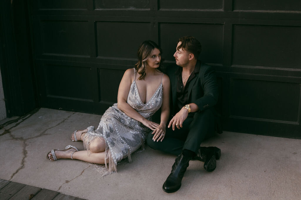 Engagement pictures photoshoot by Morgan Ashley Lynn Photography, moody aesthetic with bride and groom sitting on concrete floor in front of modern black garage door looking at eachother, the bride's arms resting on the groom's leg