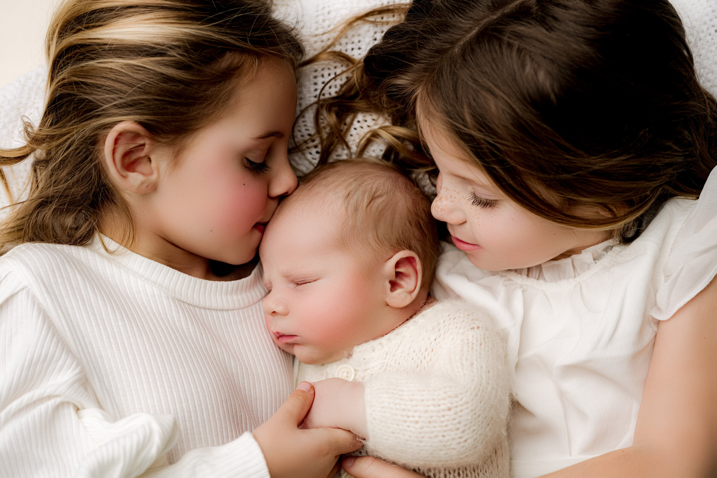 Two young girls gently embrace a sleeping baby in the center. The girls wear white outfits, their hair brushed aside, showing tender expressions. The baby, snug in a knitted white outfit, is peacefully nestled between them—a perfect moment captured by a Waxhaw newborn photographer.