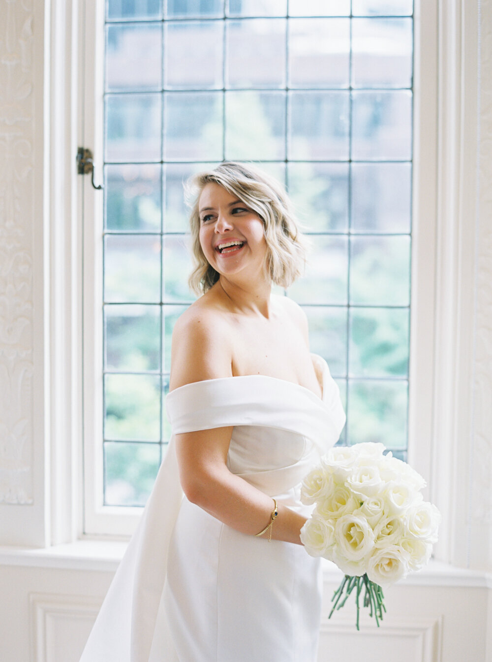 portrait of bride in her off the shoulder gown with short blonde bob haircut and white long stem roses in front of large window
