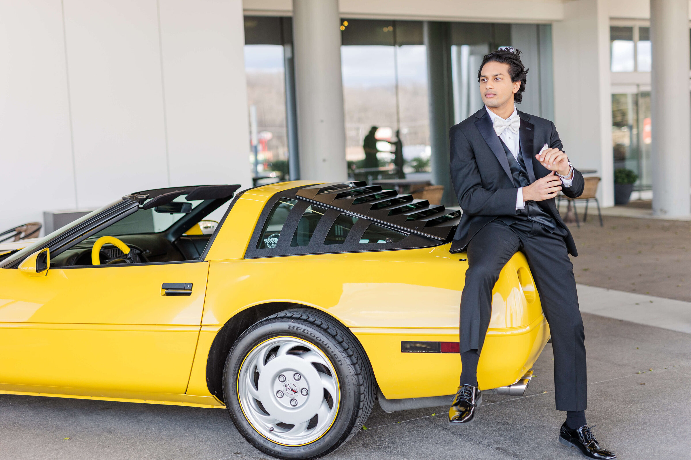 A groom sits on a yellow luxury car as he looks off into the distance