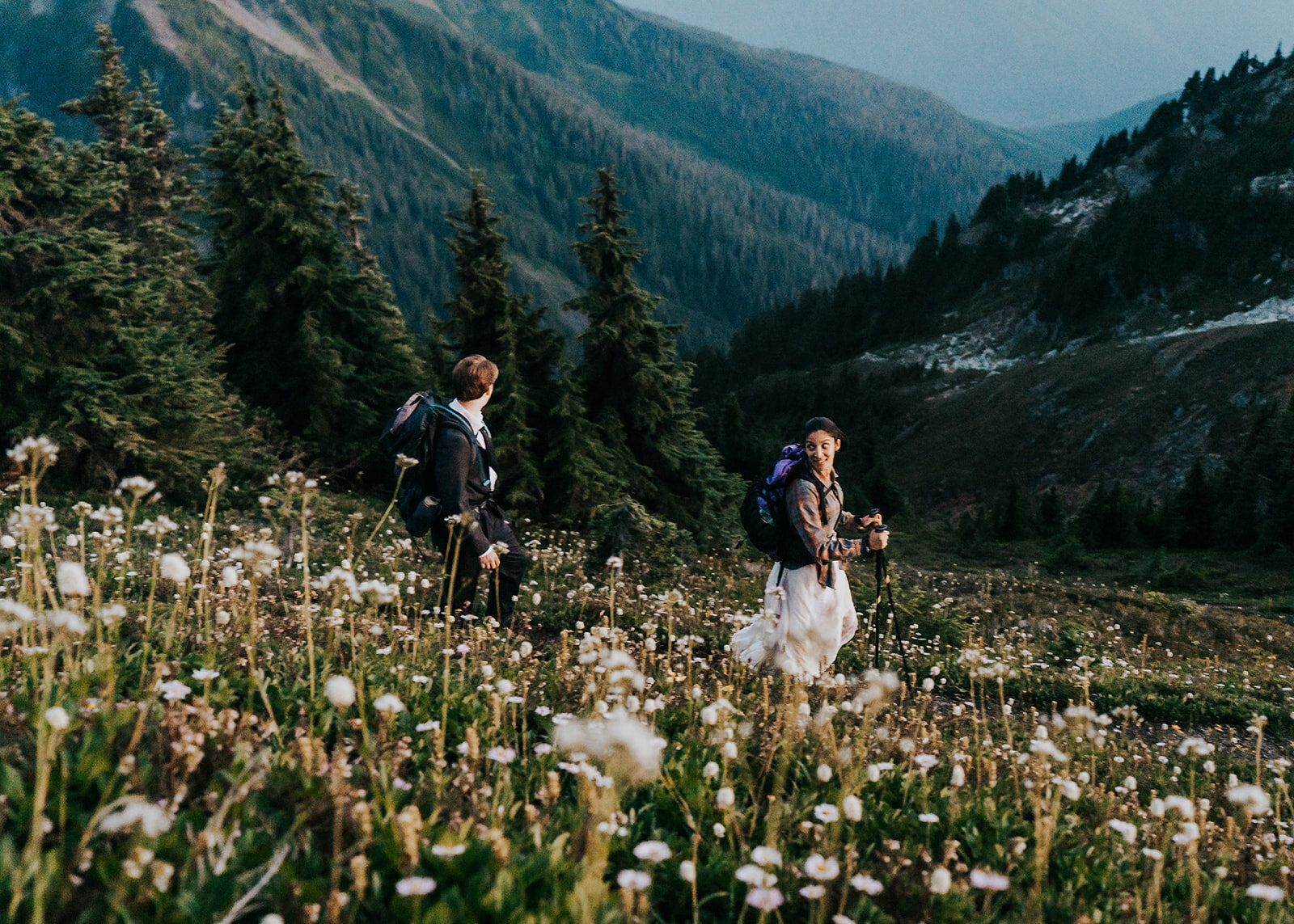 A bride and groom, in their wedding attire hike down a switchback. The bride glaces back at her groom smiling. Their adventure elopement photographers shoot from a switchback above them, showing off the mountains behind them and painting the foreground with wildflowers.
