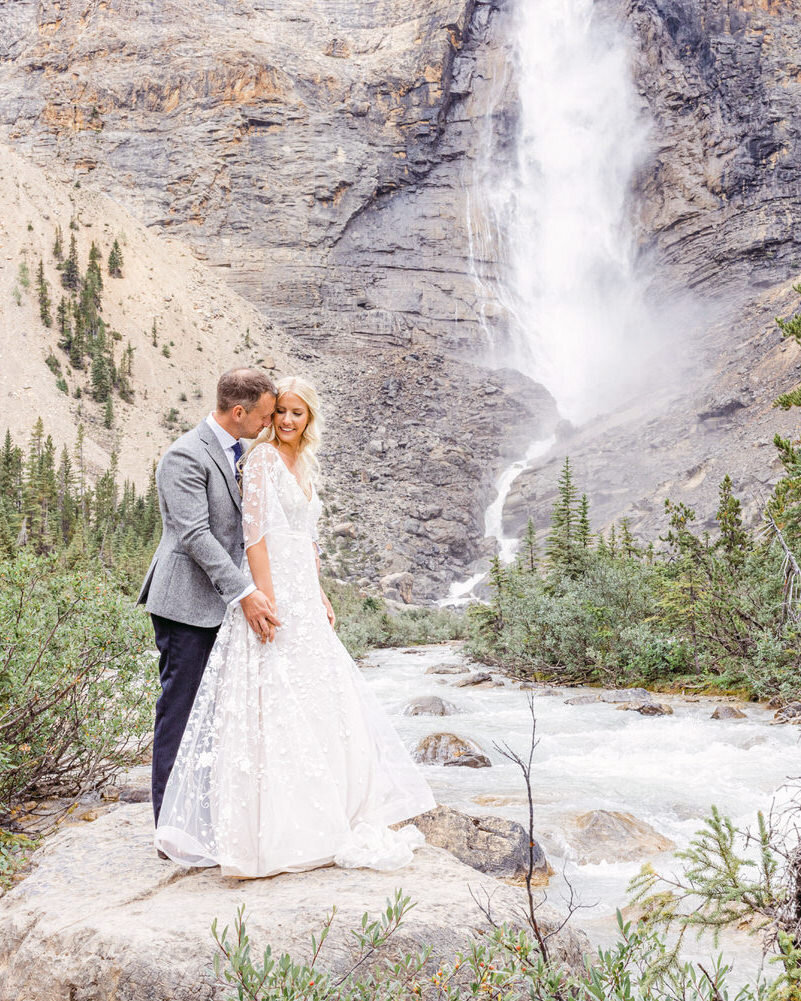 This stunning image captures the bride and groom holding each other in front of the majestic Takakkaw Falls in Yoho National Park. The powerful waterfall and natural beauty of the surroundings create a breathtaking backdrop for this romantic moment.