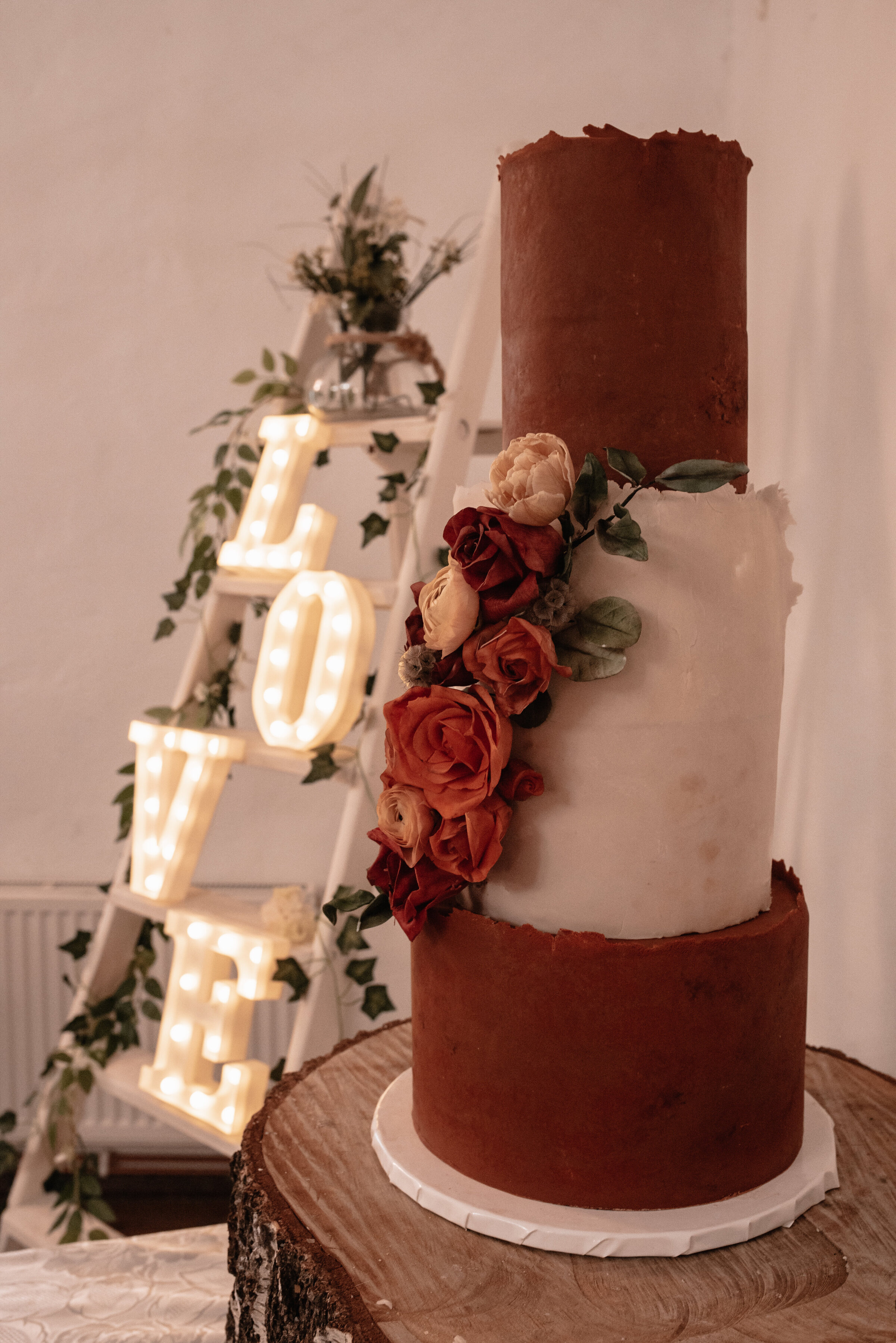 Three tier wedding cake decorated with real flowers in front of wedding ladder with light up love signs