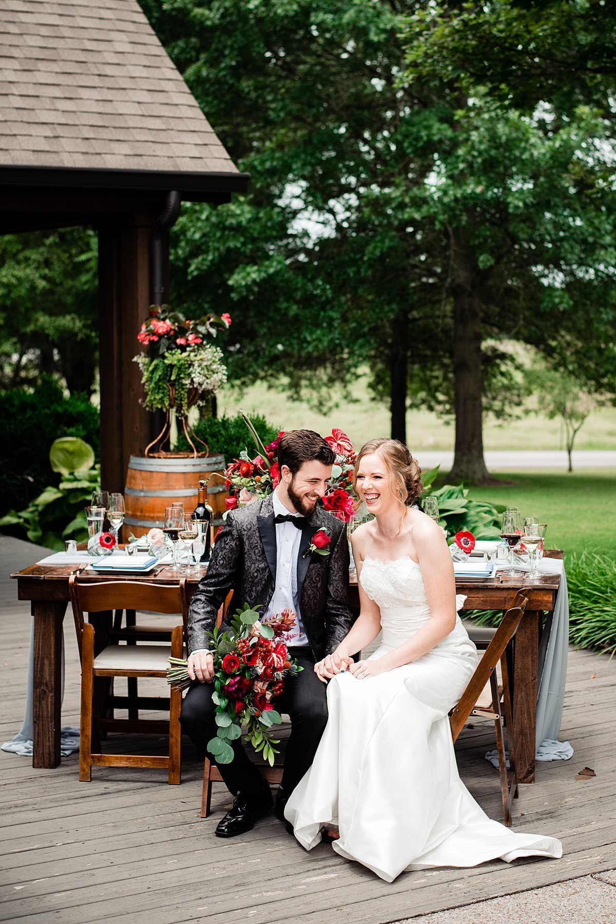 Groom wearing black tux with baroque jacket and bowtie, sitting beside his wife at an outdoor reception at Arrington Vineyards surrounded by red flowers