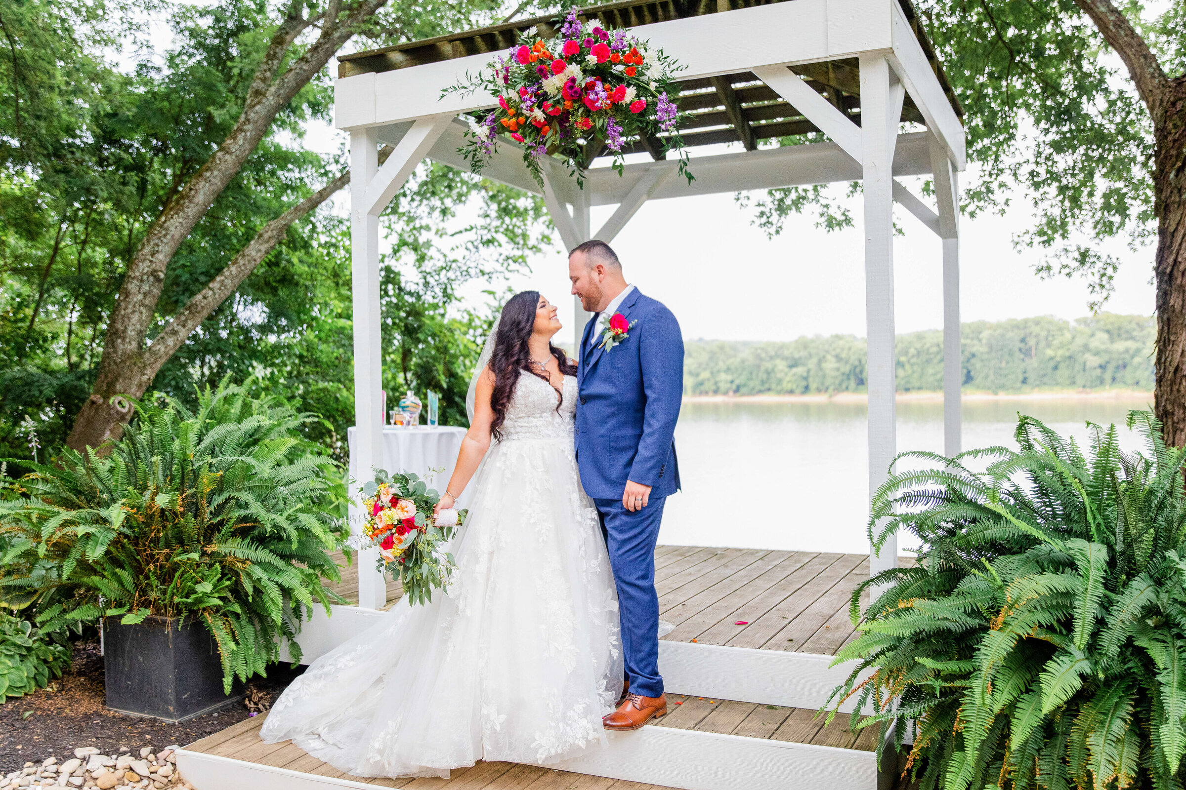 A bride and groom stand at their ceremony location, a pergola. The bride is carrying a white and pink bouquet and is on the lower step  while the groom, in a blue suit, is above her, smiling down at her.