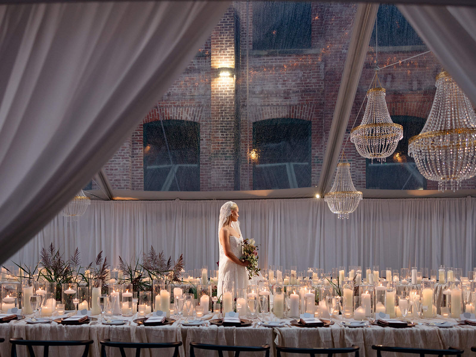 Bride at a long candlelit table beneath crystal chandeliers in a draped tent, planned by Briggs & Co.