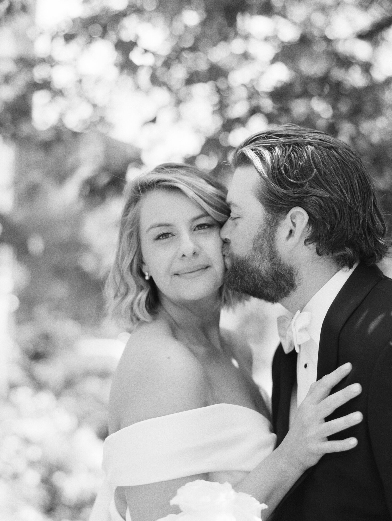 classic black and white film photo of a groom kissing his bride on the cheek