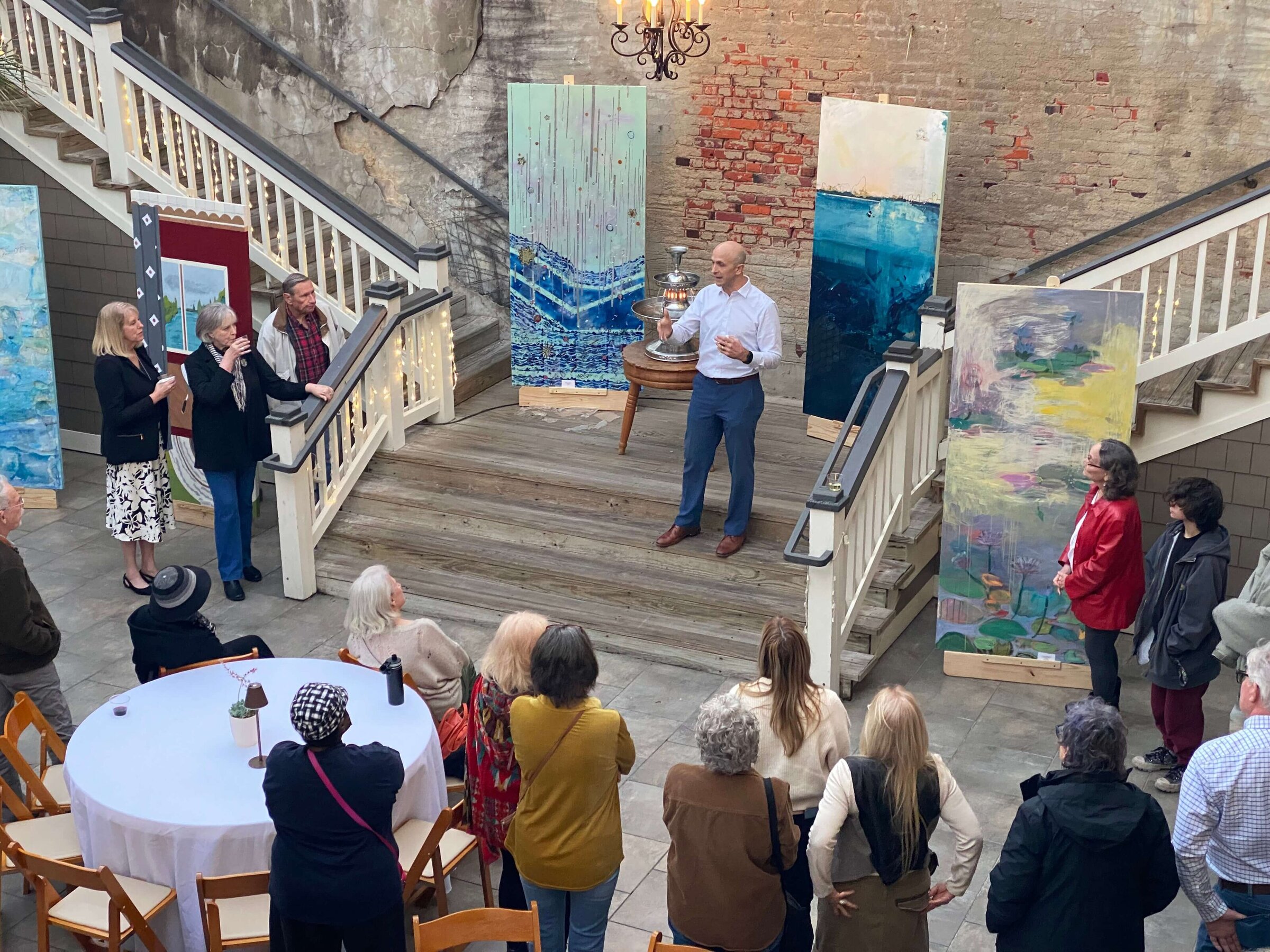 A man stands on stairs speaking to a gathered crowd at an art exhibit on the stairs in an open couryard. Large paintings are displayed around him, and people listen and observe from below.