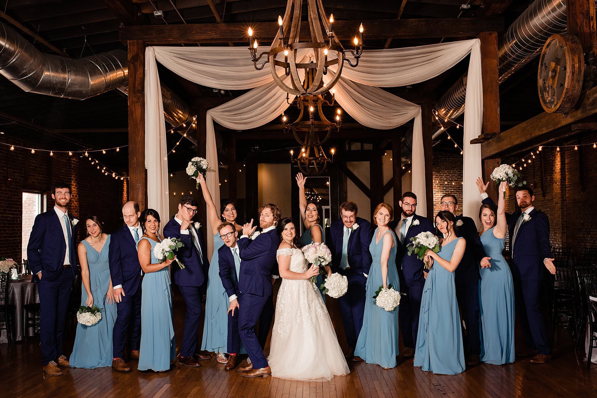 Wedding party wearing shade of blue standing together celebrating the newlyweds inside the ballroom at Cannery ONE with draping and chandeliers above